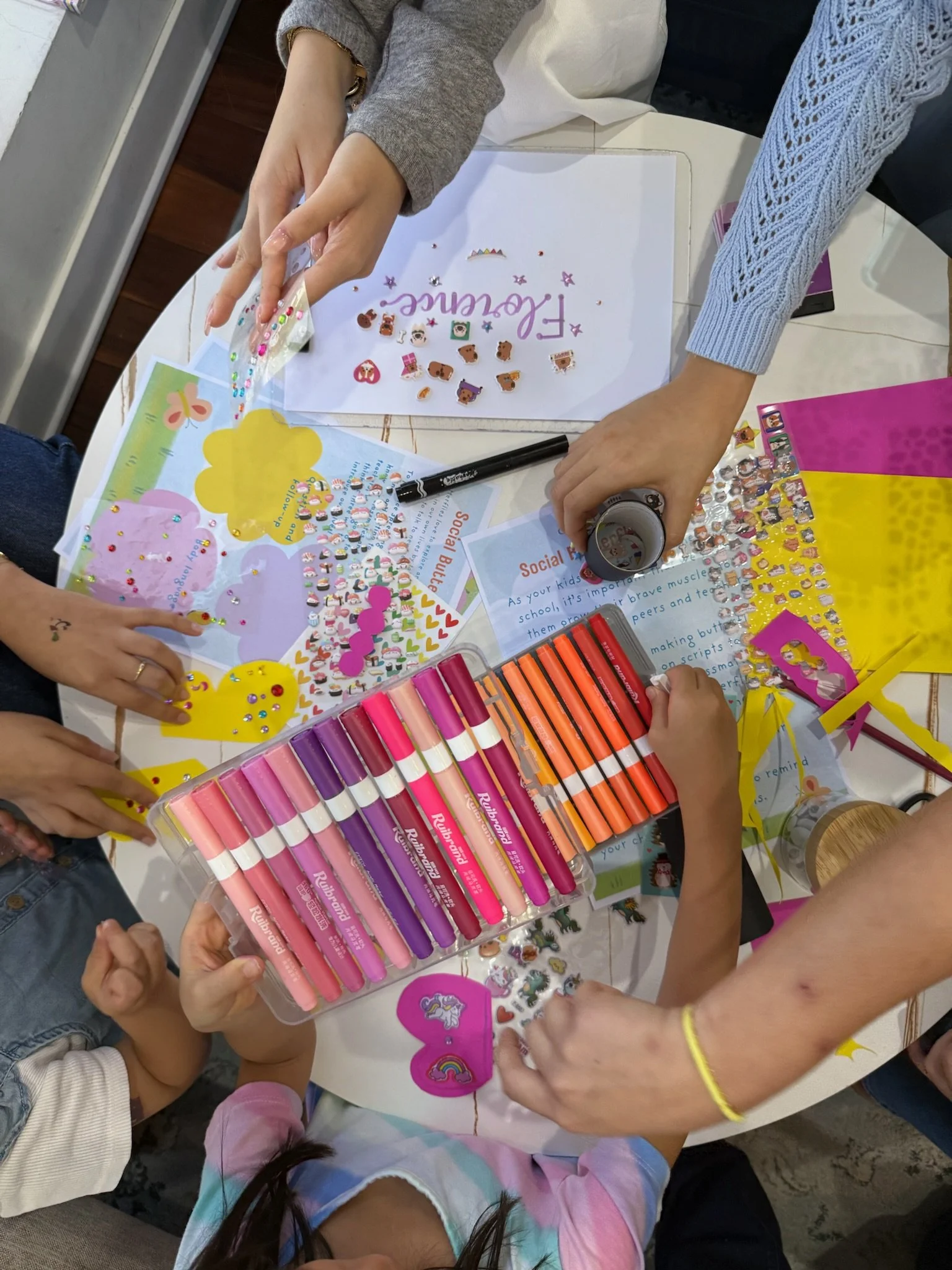 Children and adults decorating paper with colorful stickers, markers, and stickers, including unicorn and rainbow motifs. The table is filled with craft supplies and decorated papers.