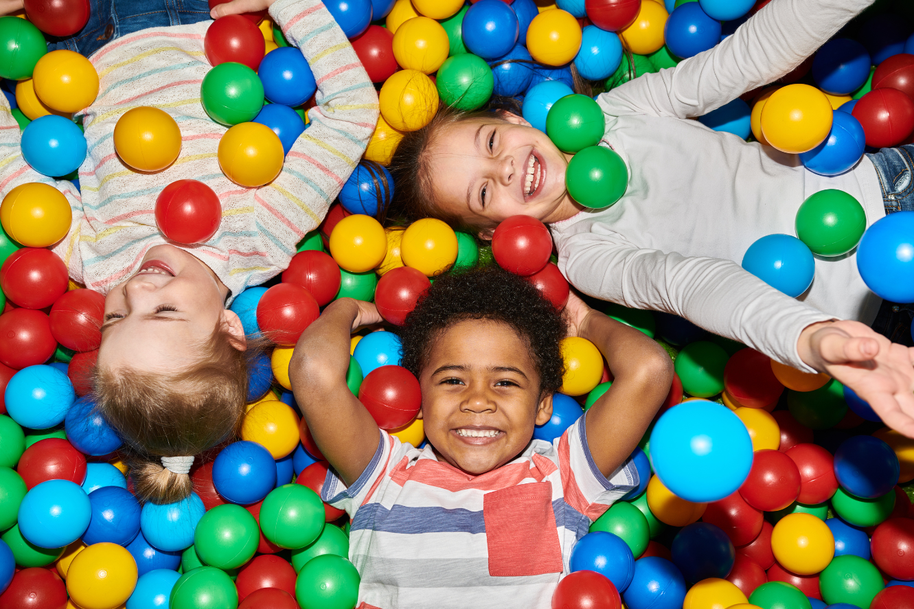 Three children lying in a ball pit smiling at the camera