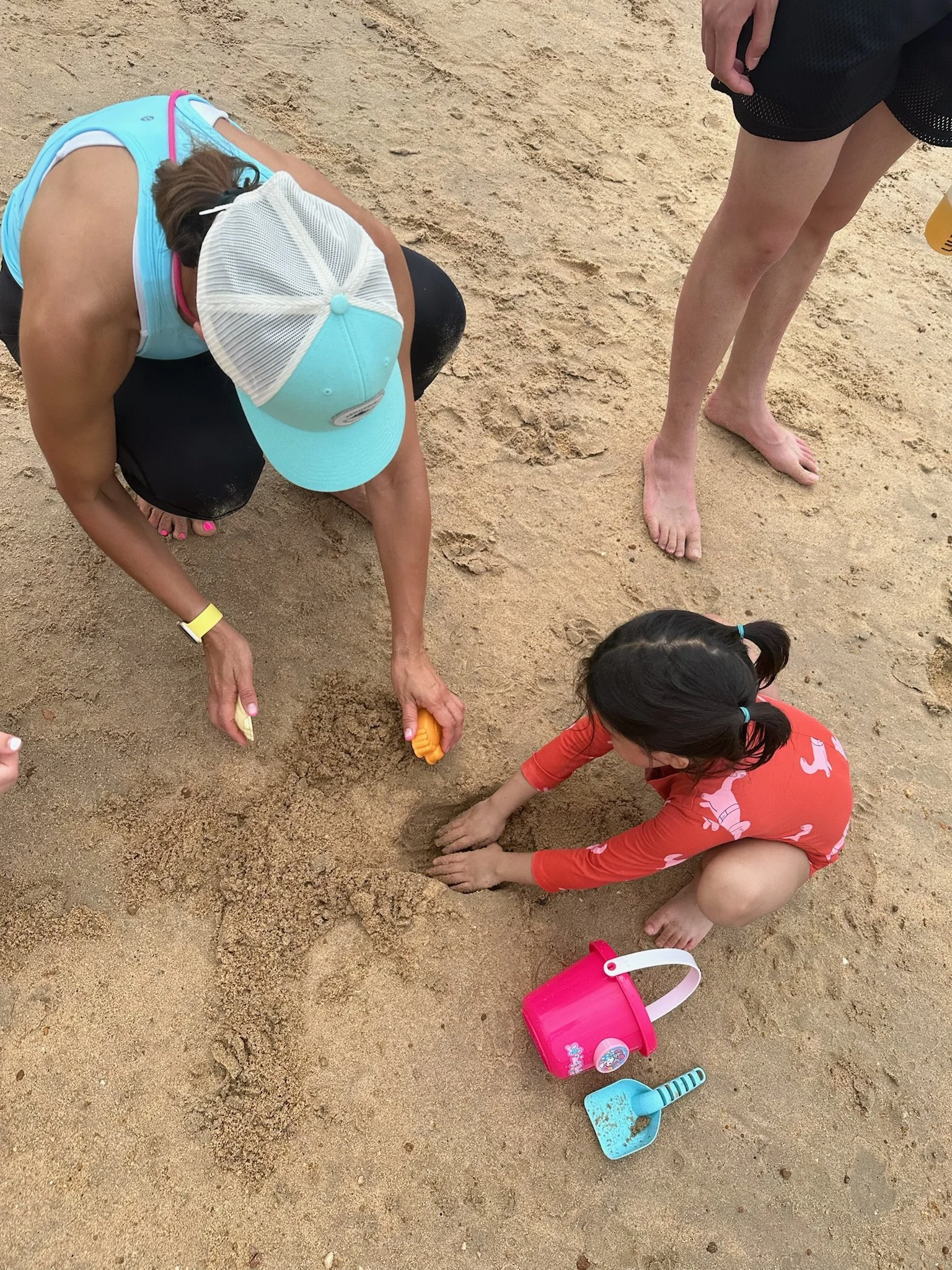 A woman and a young girl playing in the sand on the beach. The woman is crouched down, digging with a small orange shovel, and the girl is sitting and digging with her hands. There are sand toys, including a pink bucket and blue shovel, on the ground