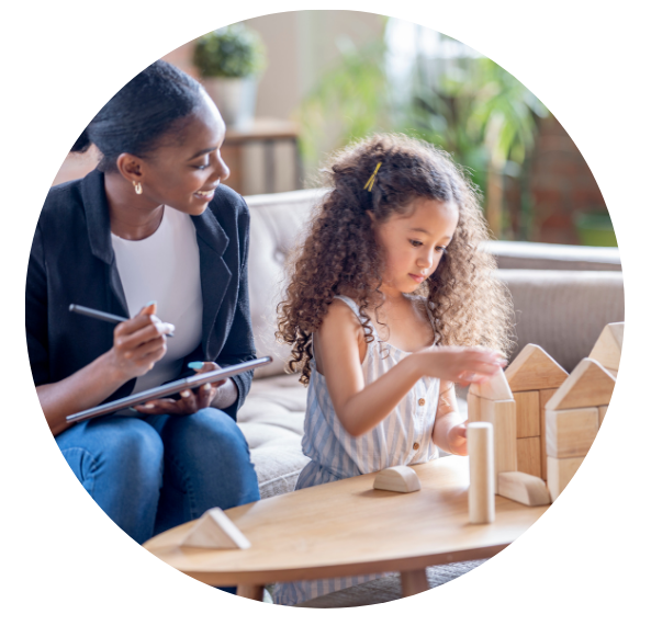 A therapist with a notepad is speaking to a young child. The therapist is smiling softly while the child builds a block tower.