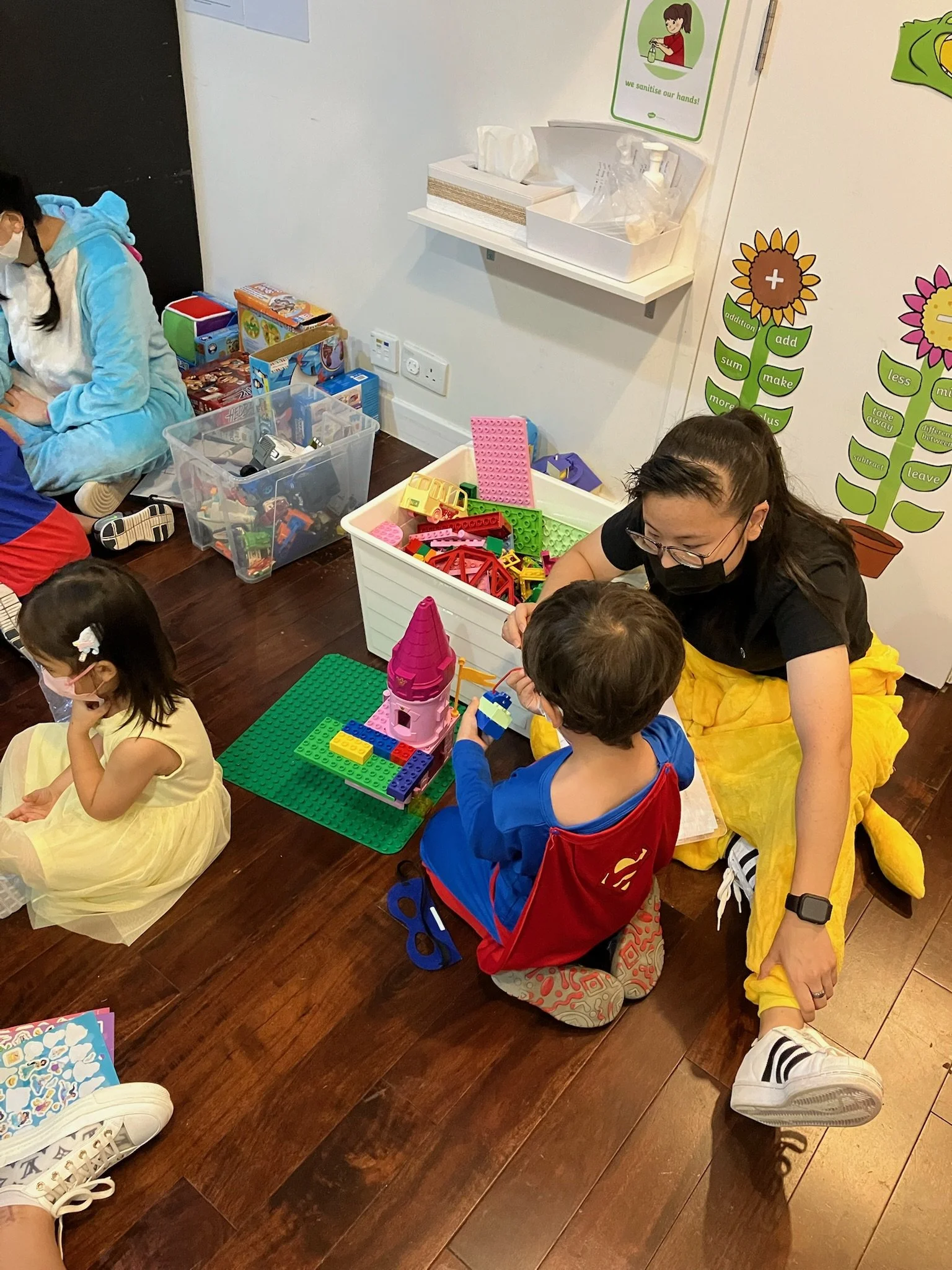 A woman dressed as Snow White sitting on the floor, helping a young boy build with colorful LEGO bricks. The boy is sitting in a red chair with a LEGO castle on a green base plate in front of him. Two children are sitting nearby, one girl in a yellow