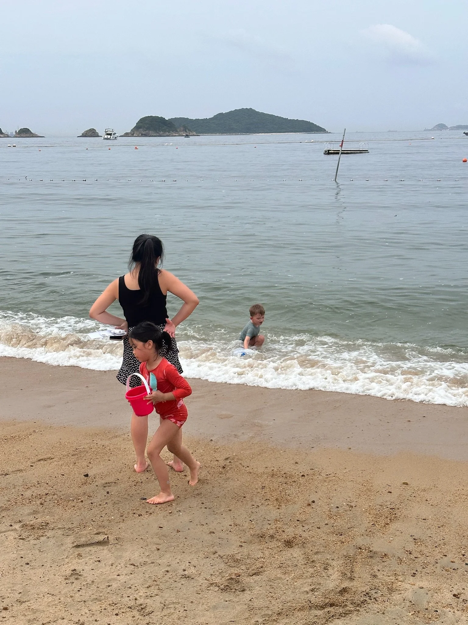 A woman and two children at the beach, one girl in a red swimsuit holding a pink bucket and a boy in the water, with islands and boats in the background.