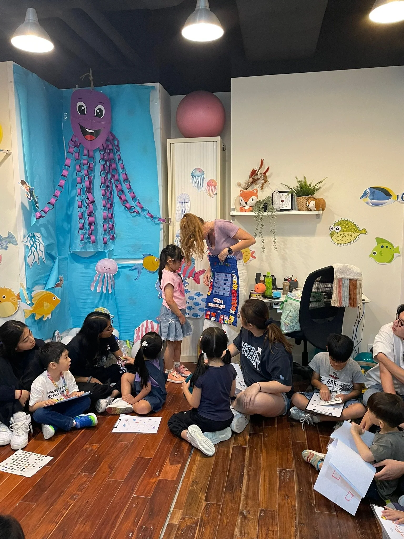 A group of children and adults sitting on a wooden floor in a room decorated with an underwater theme, including a large purple octopus, jellyfish, and fish cutouts on the walls. Two children are standing at the front, participating in a classroom ac