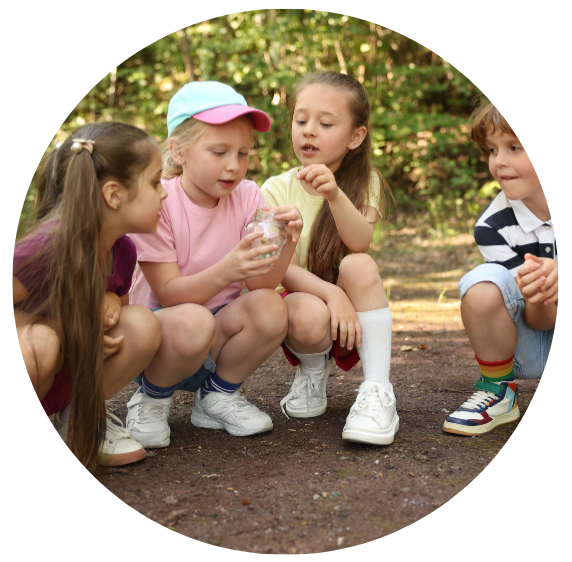 Four children squatting on the ground. They are looking at a glass jar in one of the child's hands. One child is pointing and asking a question about it.