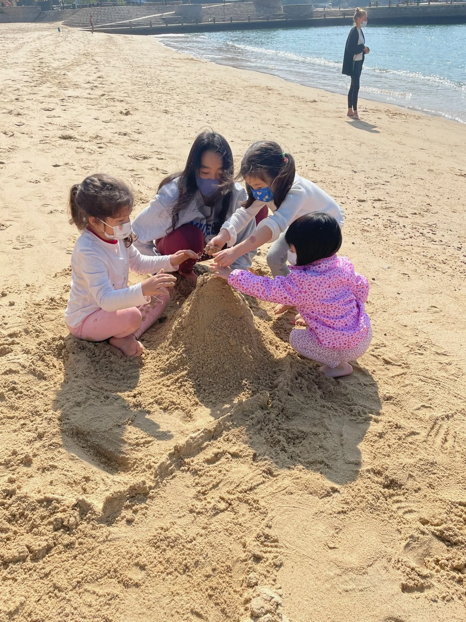 Four children building a sandcastle on the beach, all wearing masks and colorful clothing.
