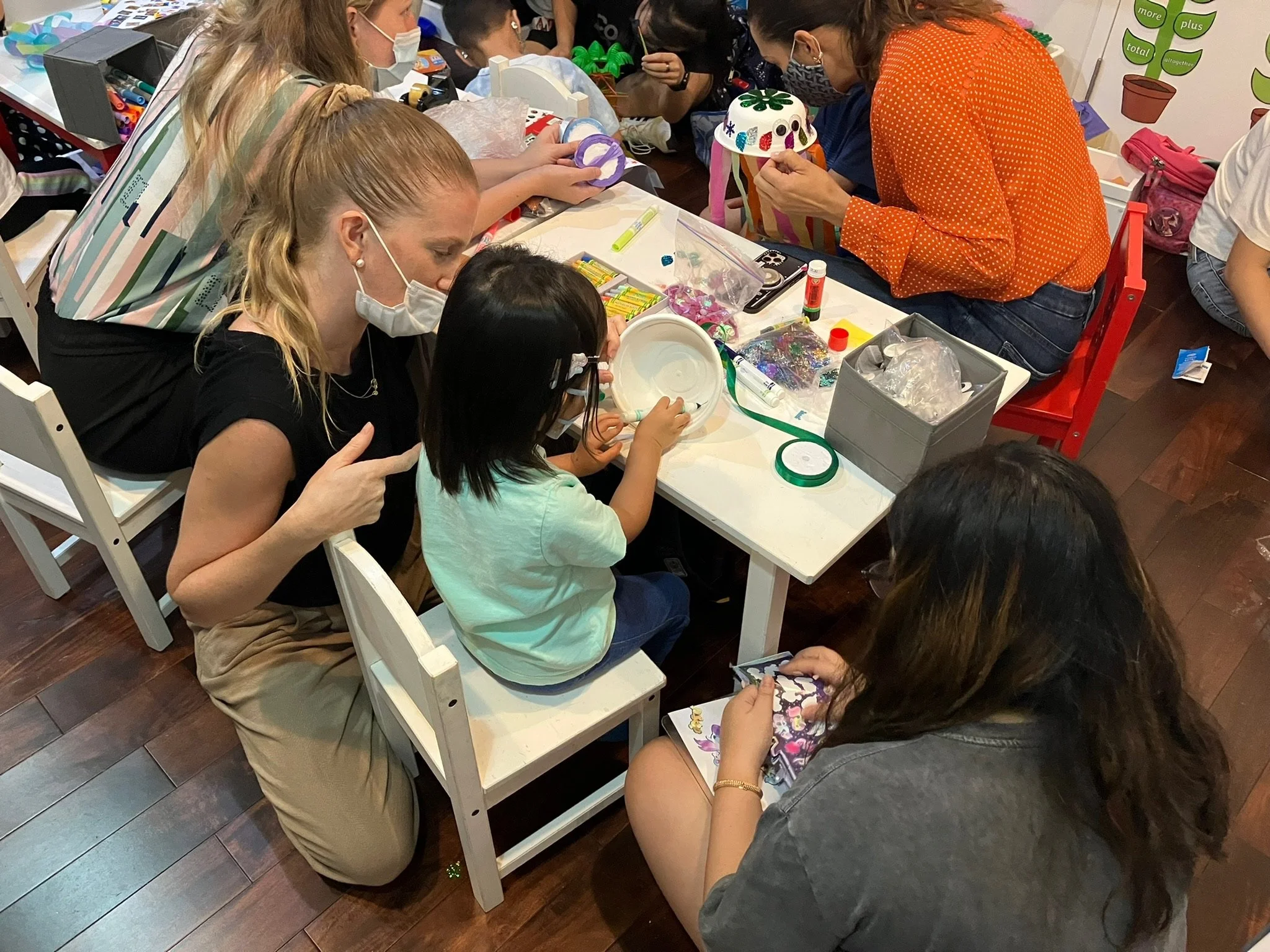 Children and adults engaged in arts and crafts activities at a table, wearing masks, with craft supplies scattered on the table, including paper, scissors, glue, and decorative items.