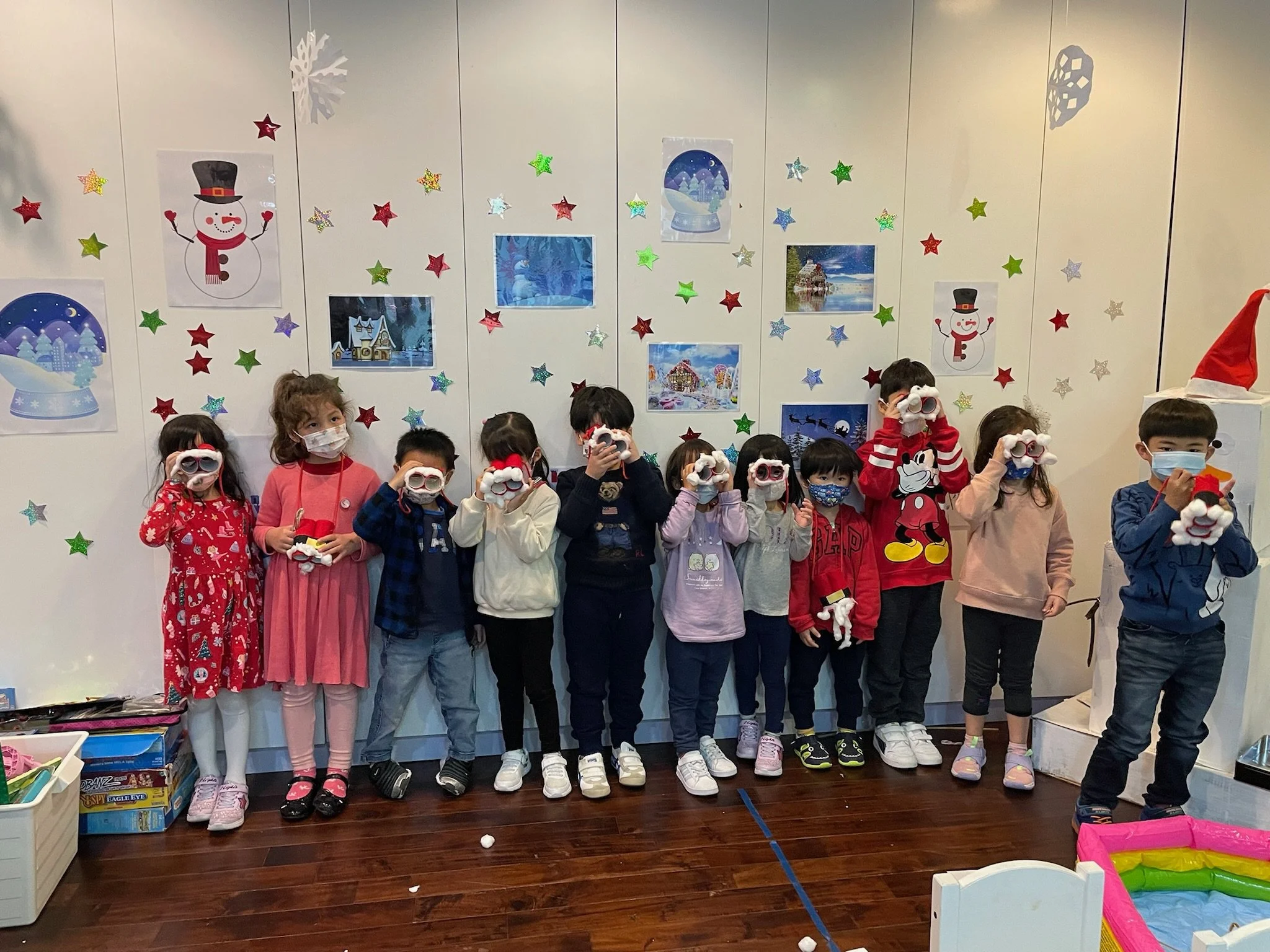 Children standing in a festive room, wearing masks and holding snowball props, with holiday decorations and colorful stars on the wall.