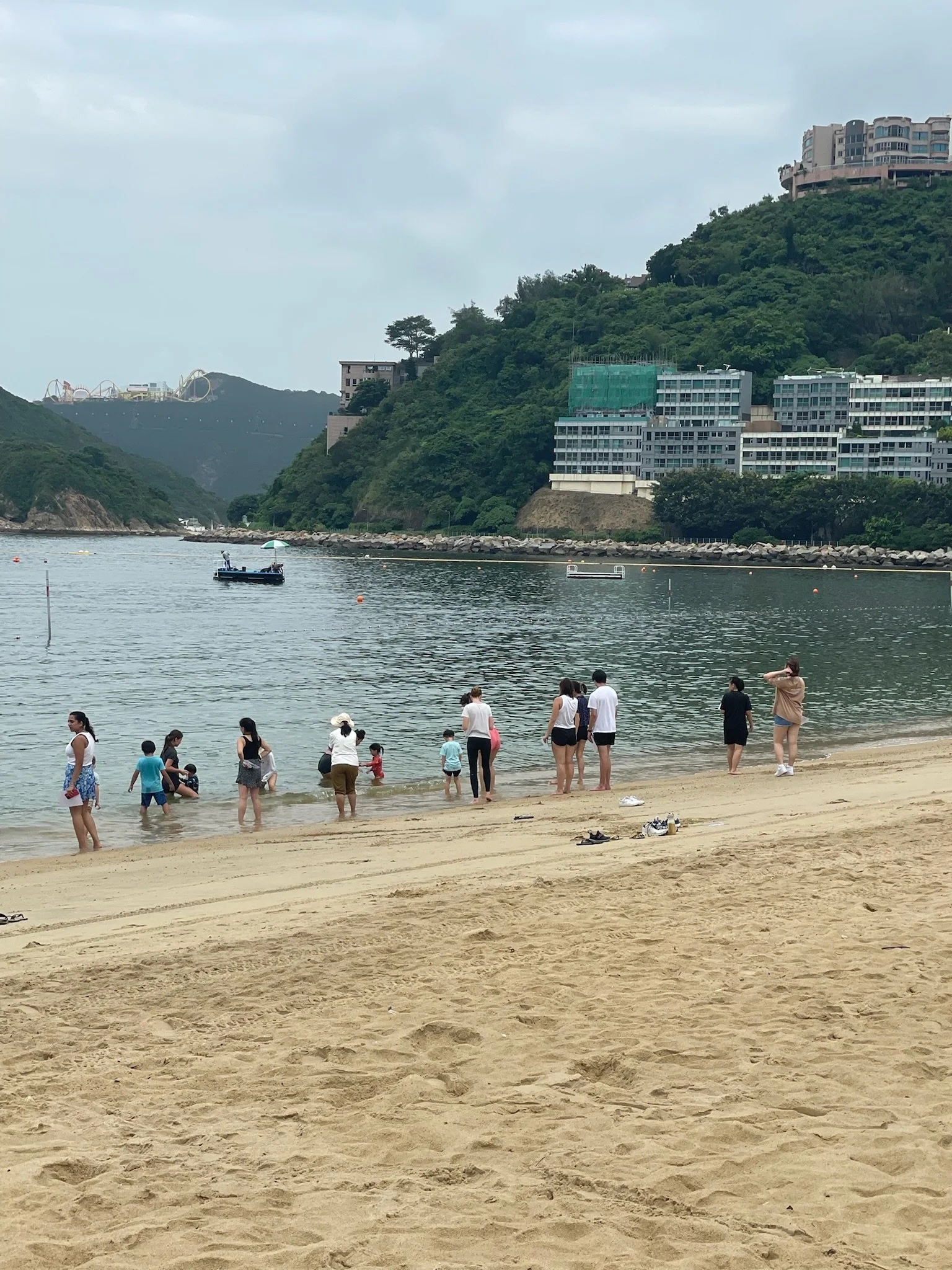 People on a sandy beach near water, with mountains and buildings in the background, some standing in the water and others on the shore.