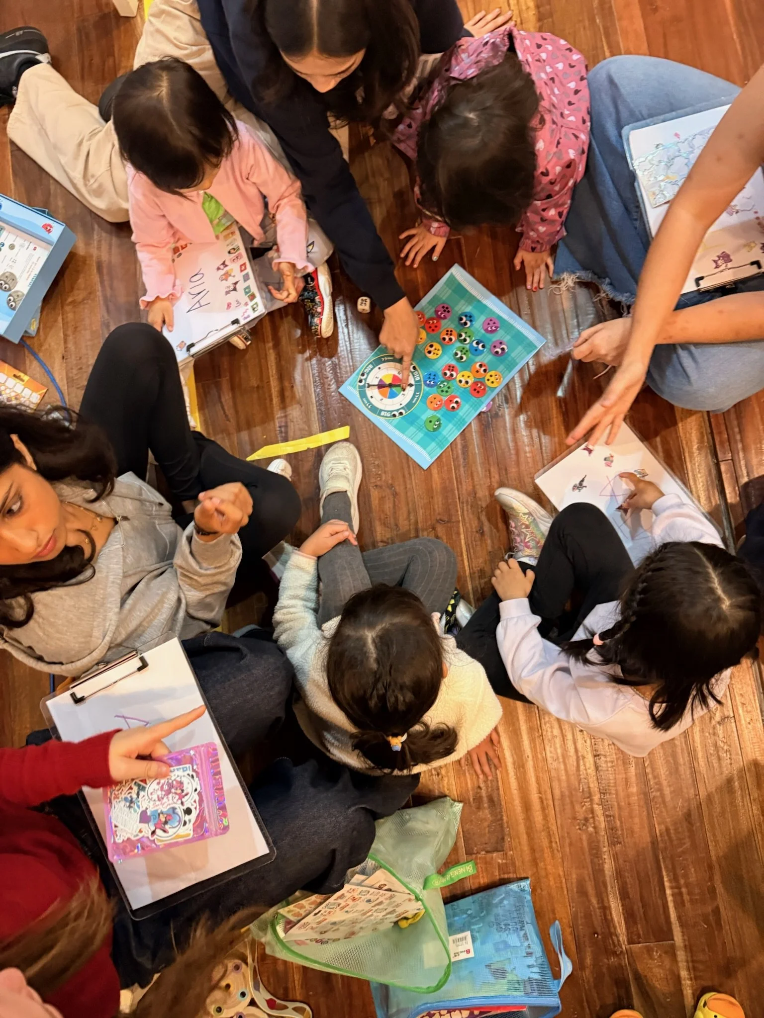 Group of children and adults sitting and lying on a wooden floor, playing board games and drawing with colorful materials.