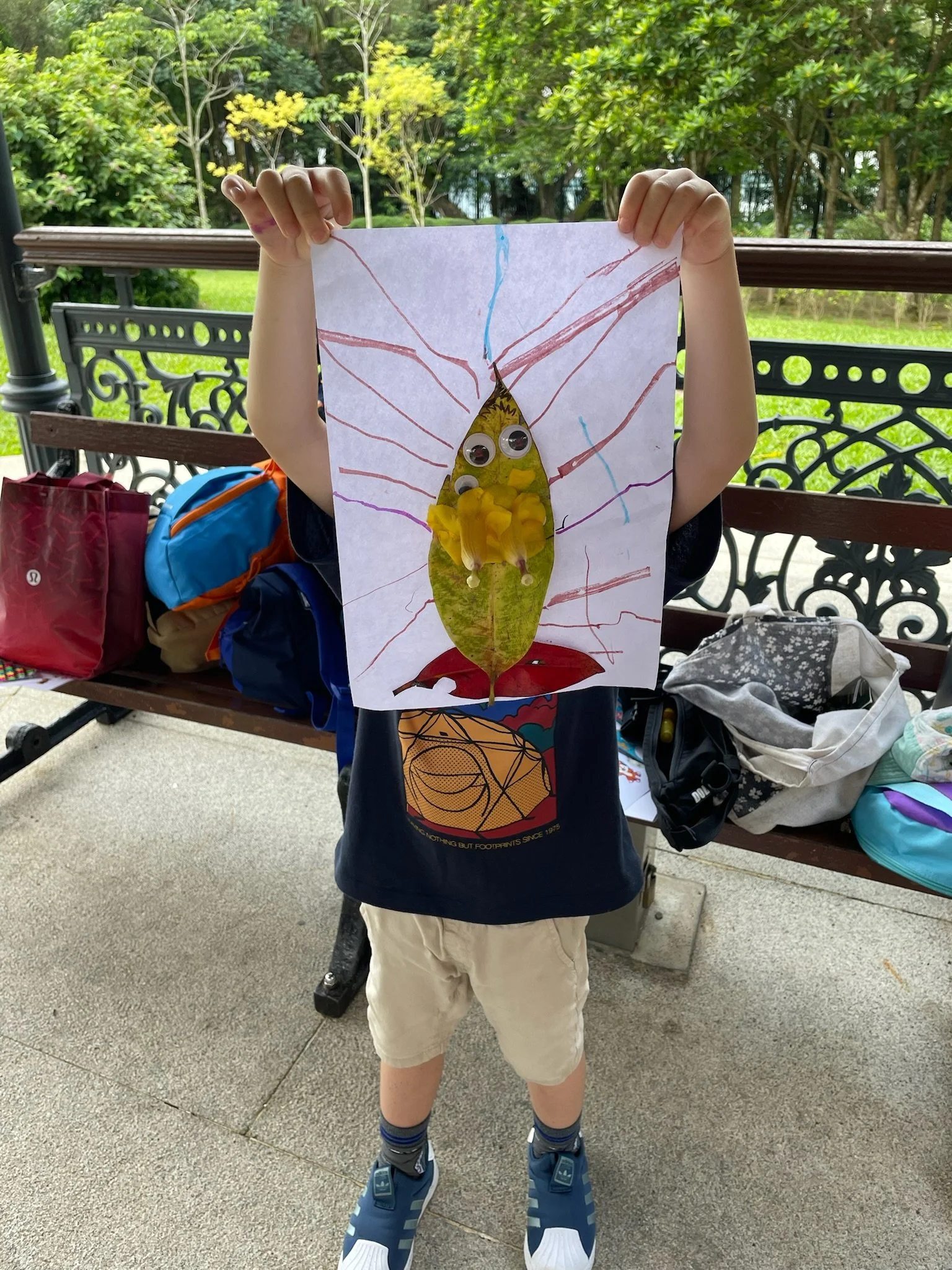 Child holding a leaf craft art with googly eyes, yellow tissue paper, and drawn red and blue lines, outdoors with a park bench and backpacks in the background.