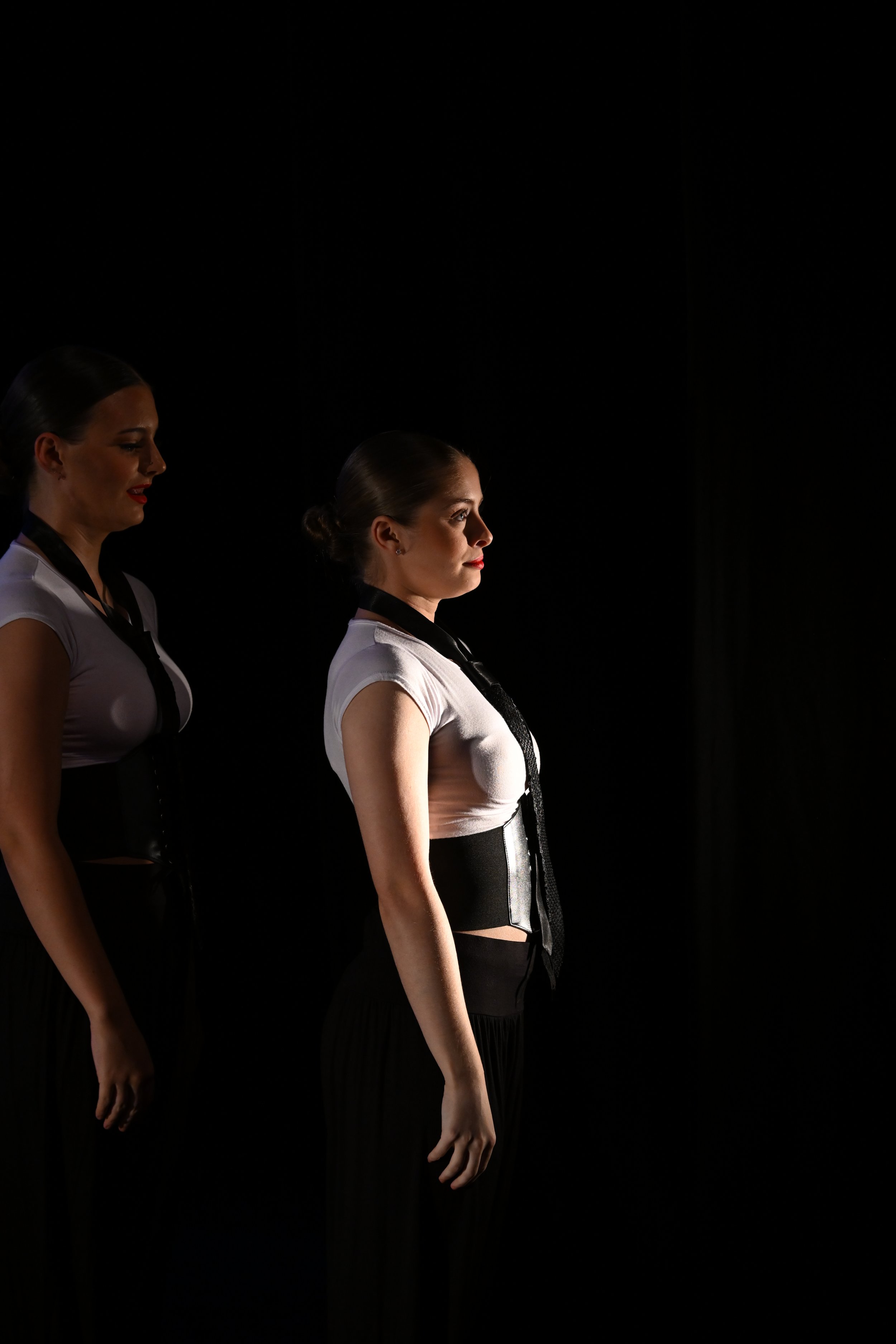 Two women standing side by side against a black background, with one woman slightly ahead of the other. They are dressed in black and white outfits with black neckties, and are looking forward with serious expressions.