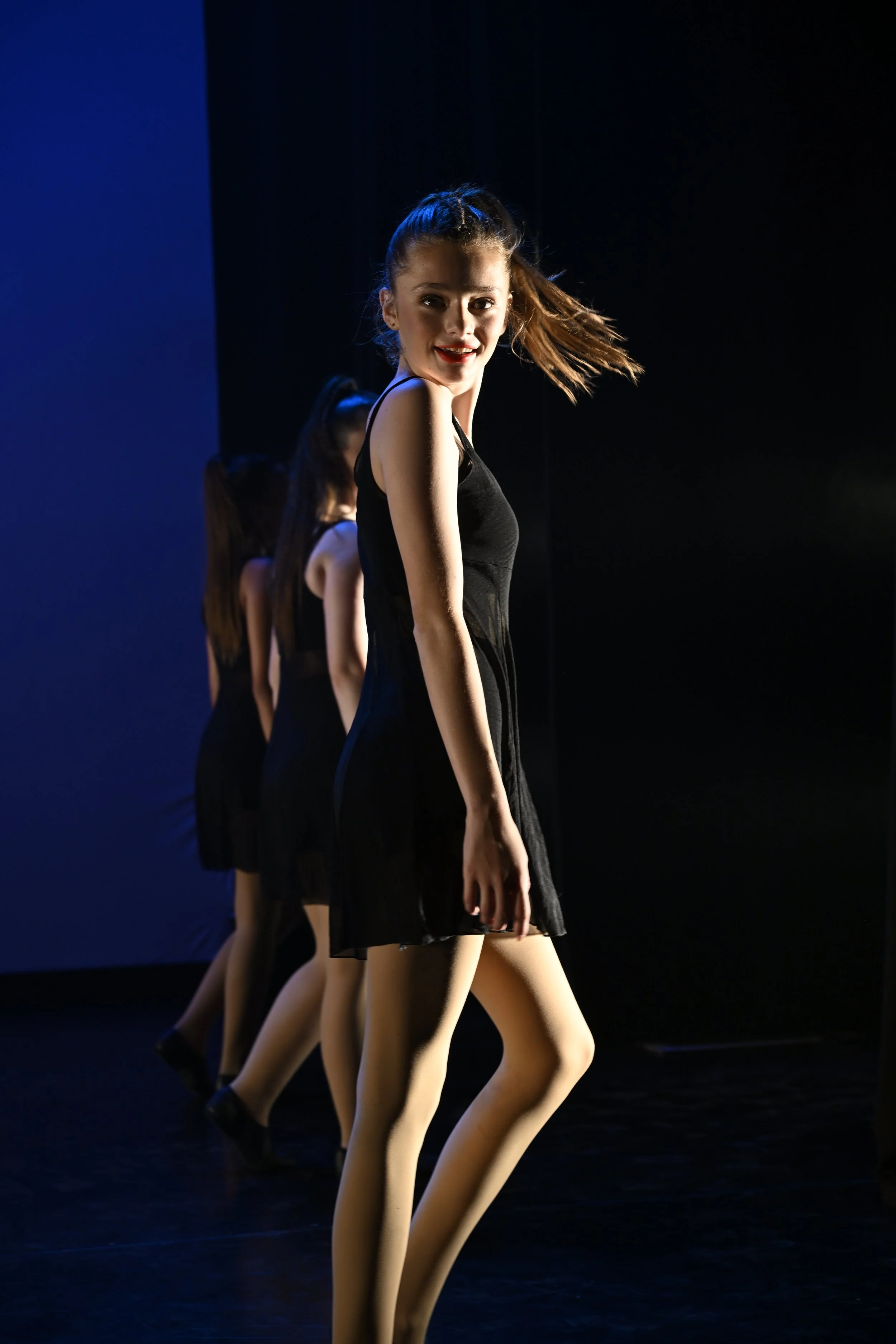 A girl in a black dress performing a dance on stage, with three other girls in the background, all in black dresses, under stage lighting.