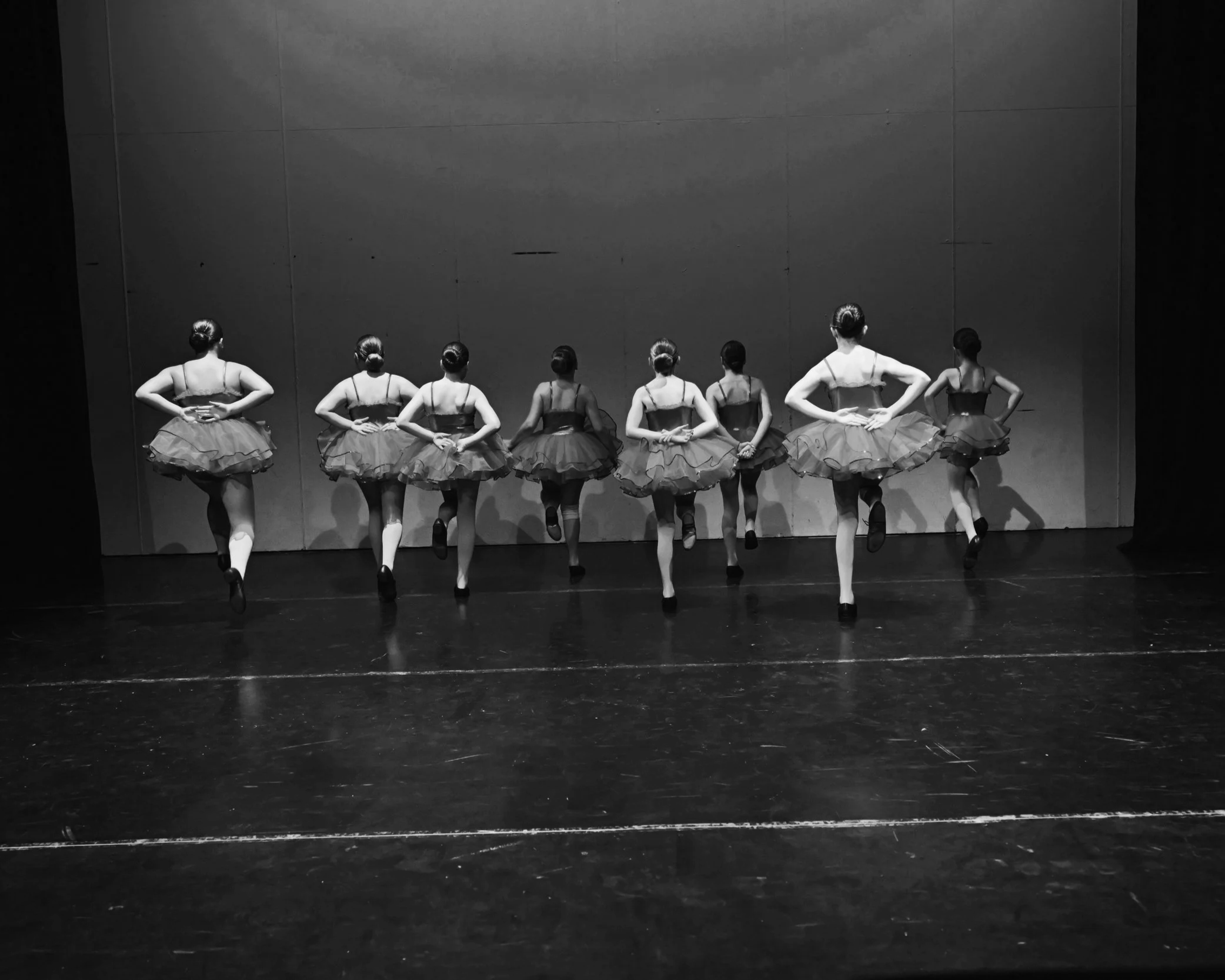 A group of ballet dancers in tutus practicing on stage, viewed from behind, in black and white.