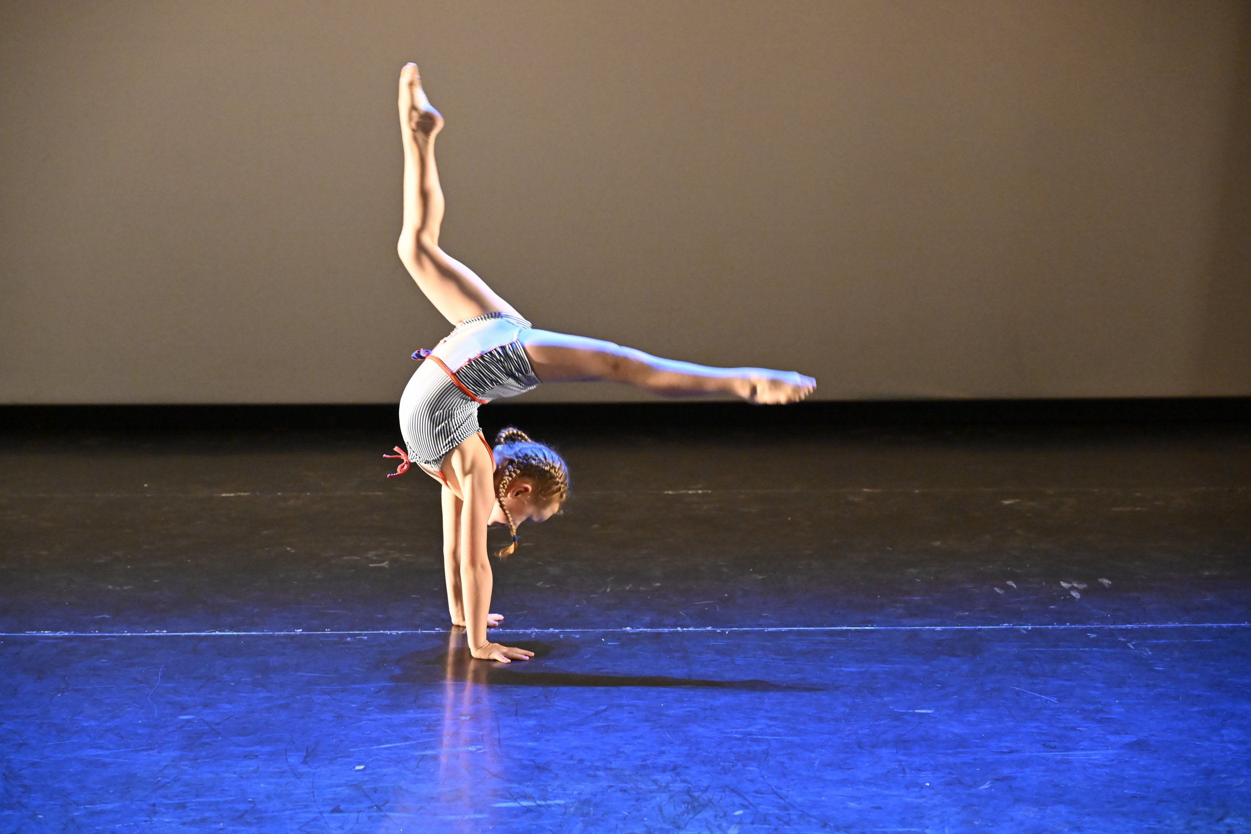 Young girl in striped outfit performing a handstand on stage with a dark background.