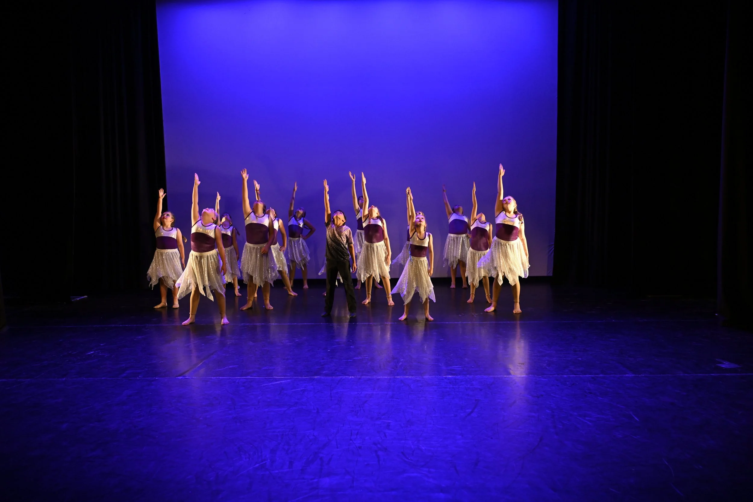 Dance performance on stage with twelve young dancers in white skirts and purple tops, led by a woman in black, under blue lighting, with black curtains on sides.