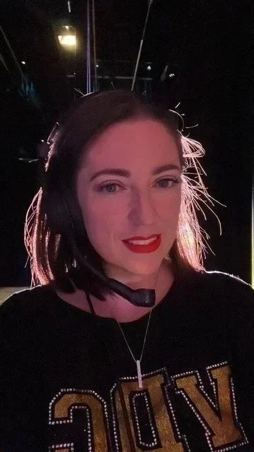 A woman with a headset, wearing a black shirt with gold sequined letters, smiling in a dimly lit environment with lights and equipment in the background.