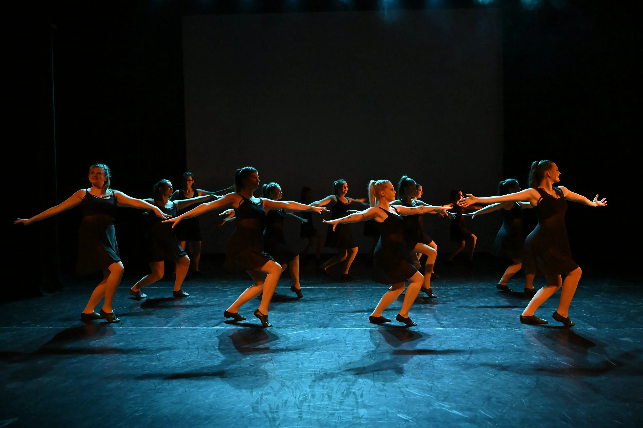 A group of female dancers performing on stage in black dresses and dance shoes, with arms extended and mid-movement, with a dark background and spotlight lighting.