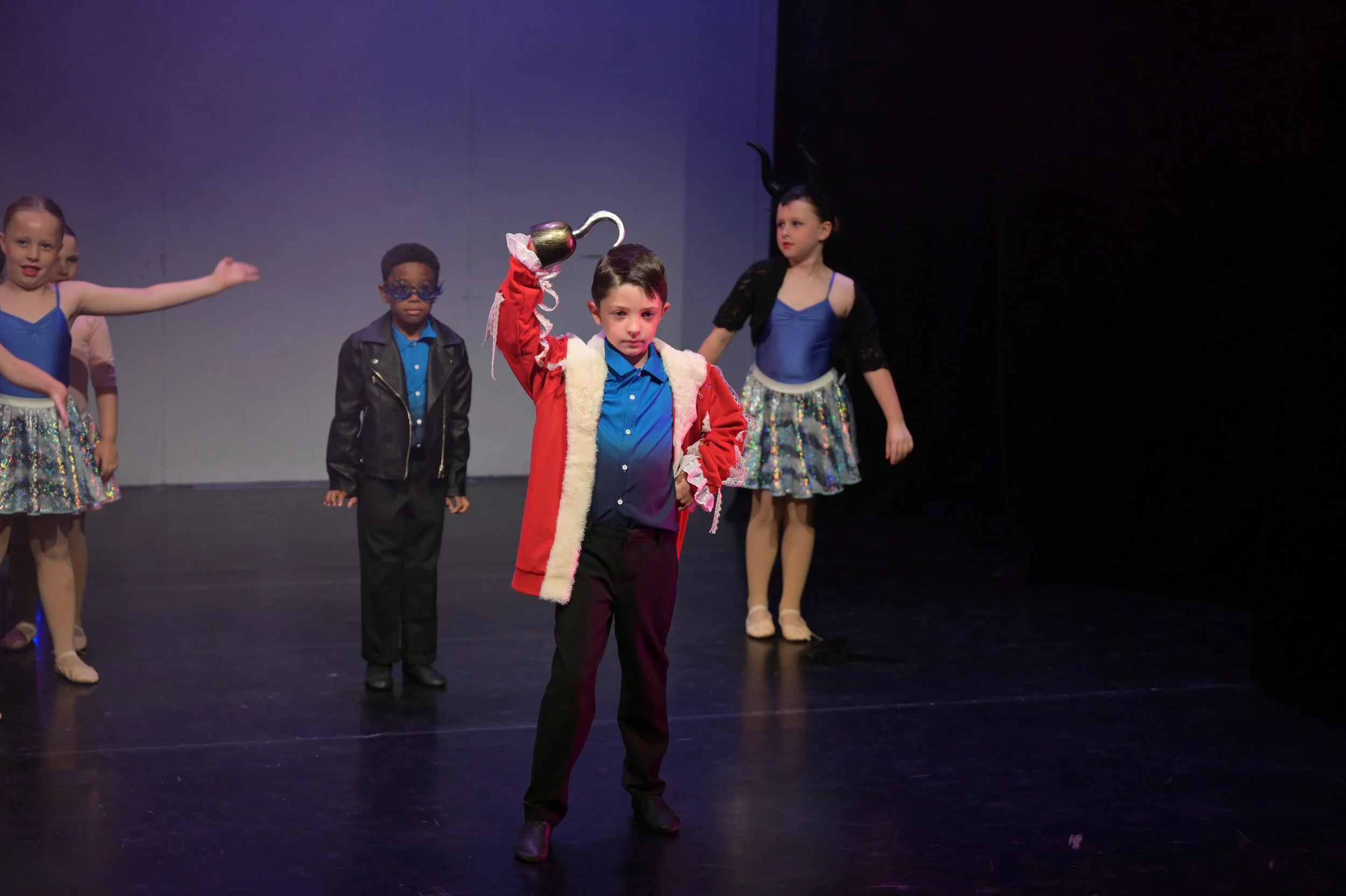 Children dressed in colorful costumes performing on stage during a dance recital, with one boy holding a silver teapot prop.