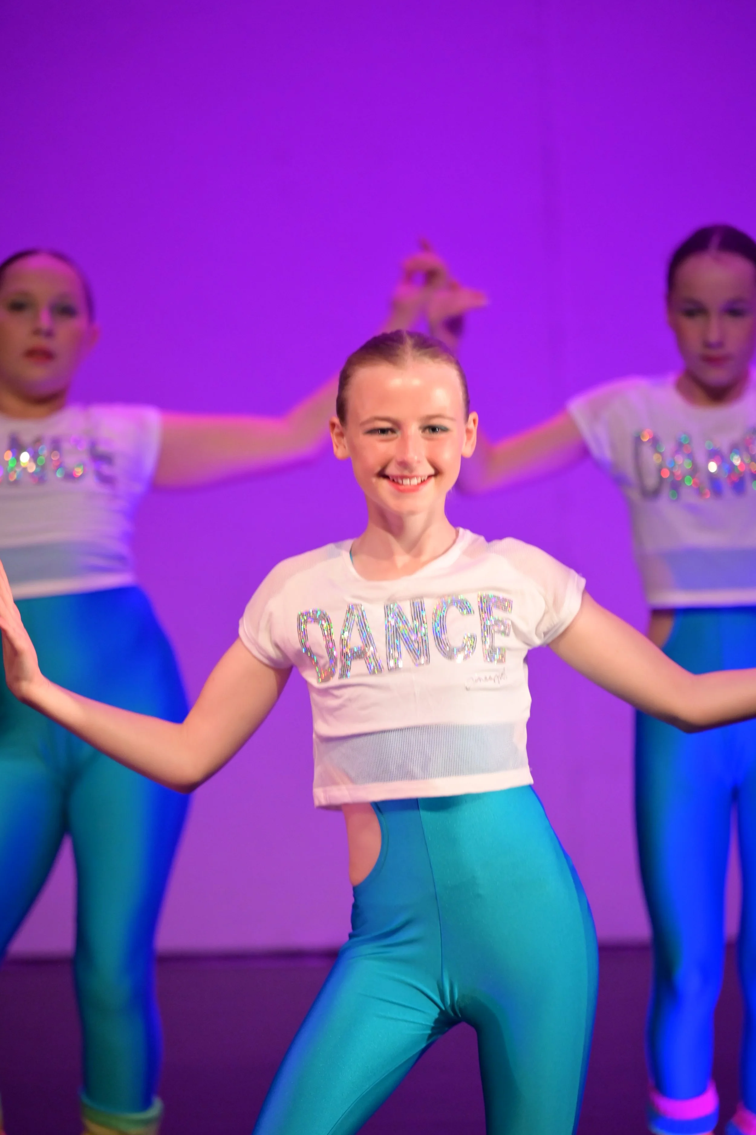 Young girl smiling and dancing in a stage performance, wearing a white t-shirt with 'DANCE' written on it in colorful rhinestones and bright blue leggings, with similar dancers in the background.