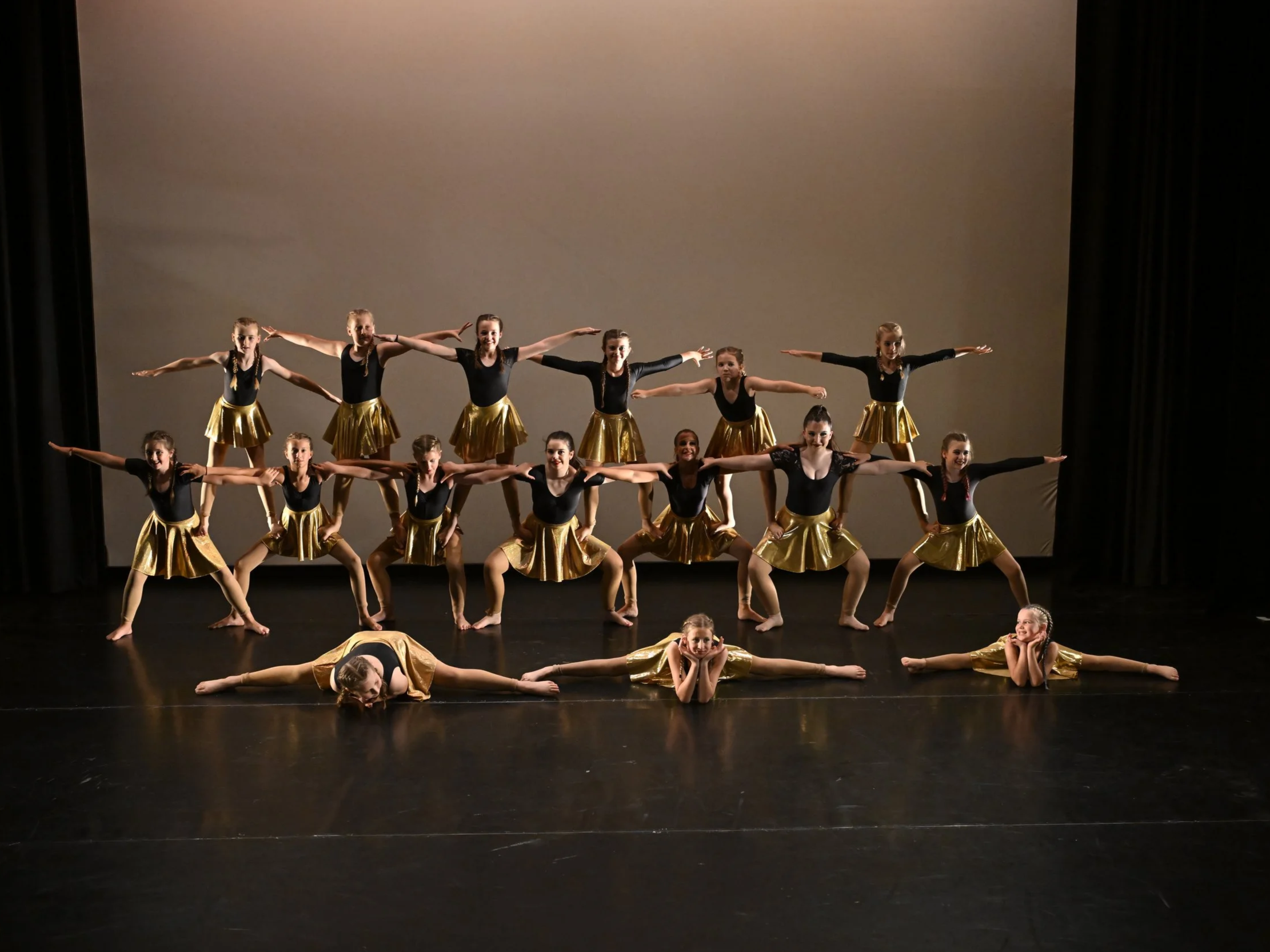 Group of young female dancers in black tops and gold skirts performing a dance routine on stage with a plain backdrop.