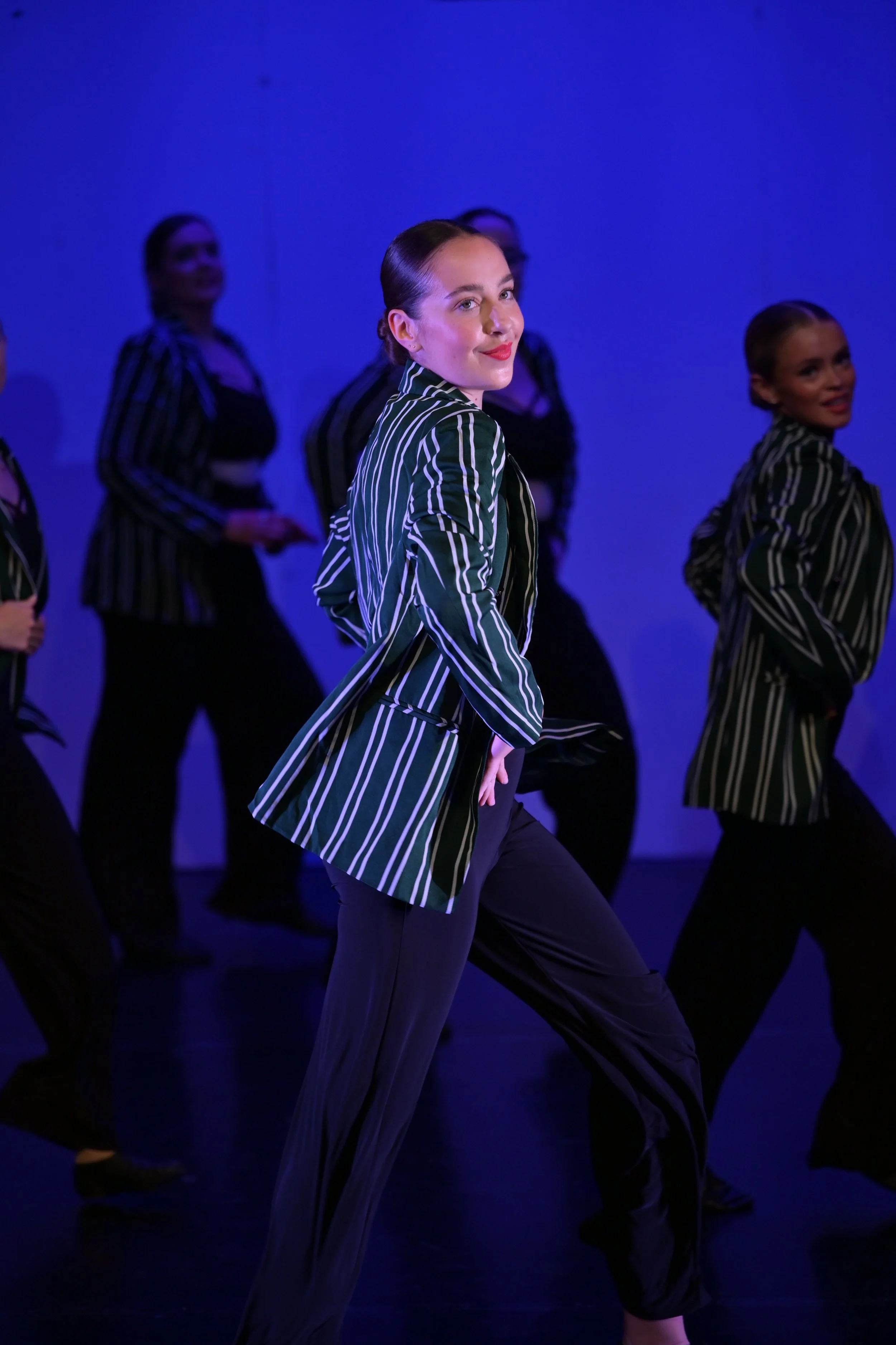 A woman in a striped blazer poses among dancers in similar outfits during a stage performance with blue lighting.