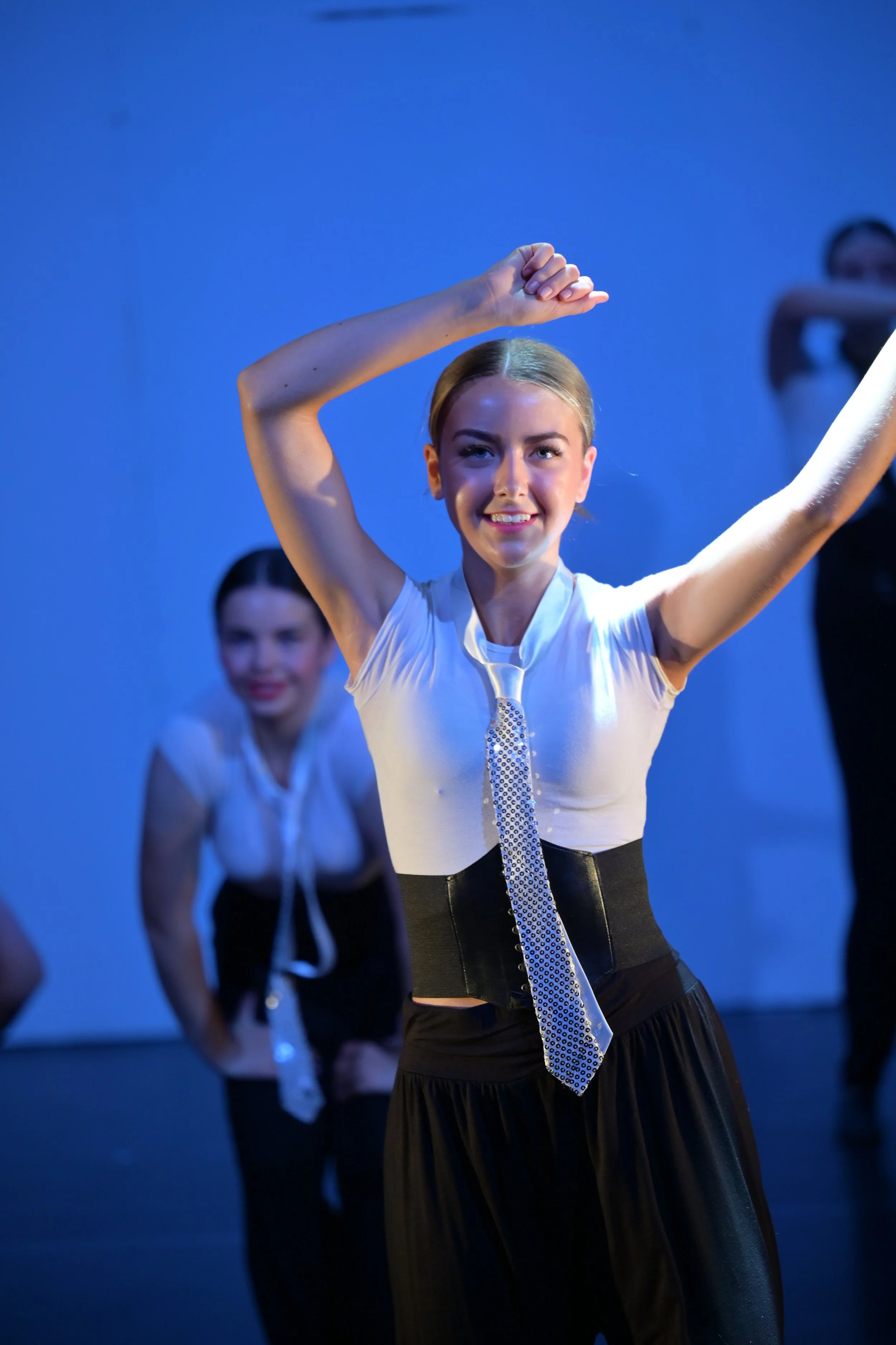 Young woman with blonde hair smiling and posing with arms raised, wearing a white top, black skirt, and a tie, with other women in similar outfits in the background on a stage with blue lighting.