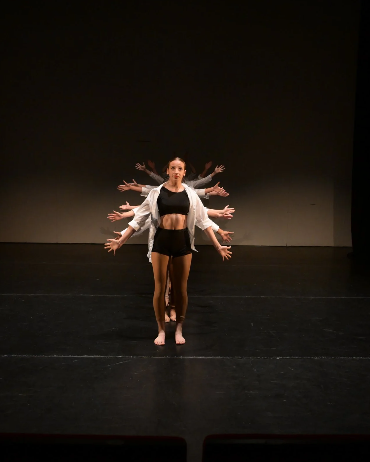 A performer standing in front, with several other performers behind her, all with arms extended outward, against a dark background, creating a layered visual effect.
