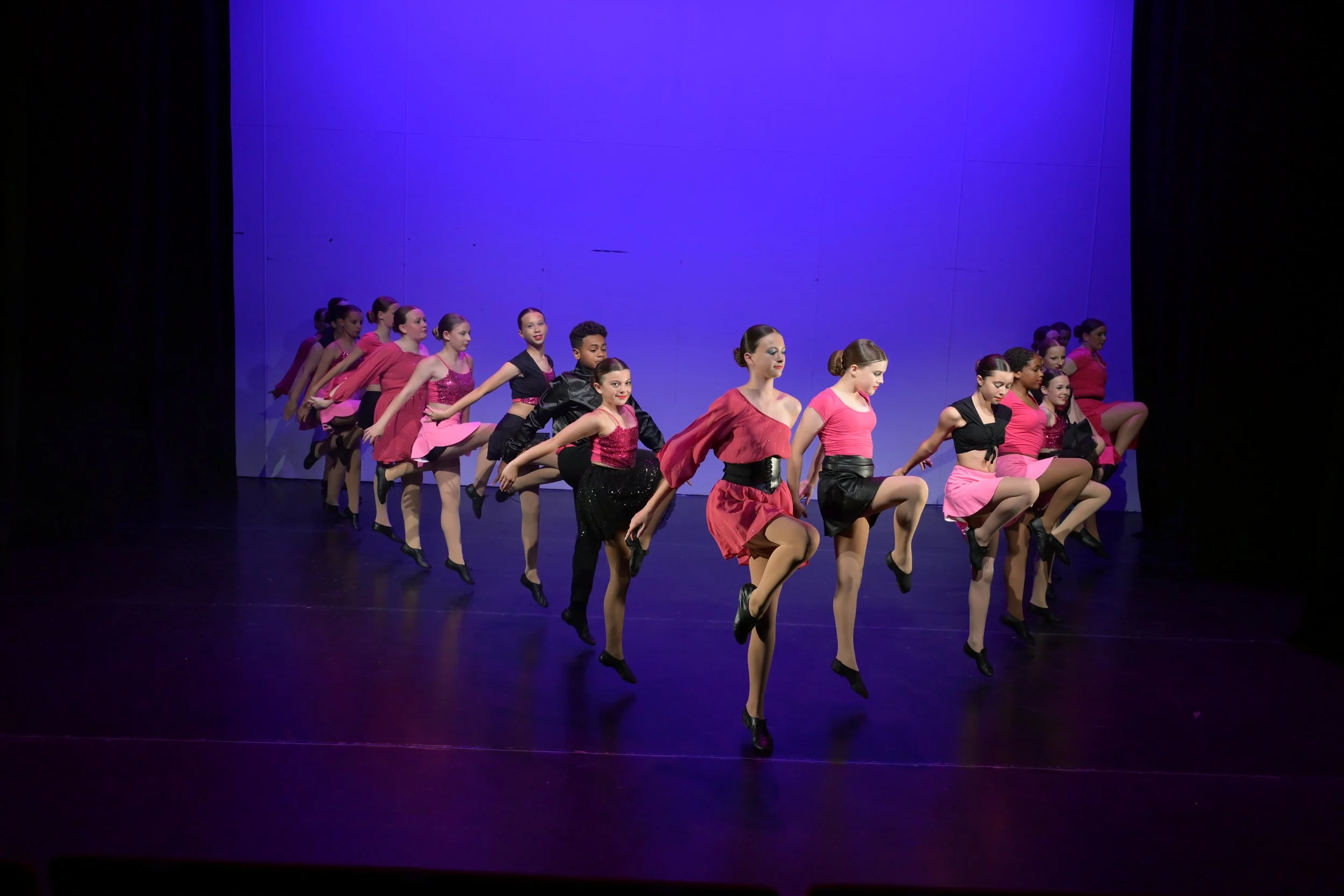 A group of female dancers performing a choreographed dance on stage, dressed in coordinated black and pink costumes, with a purple-lit backdrop.