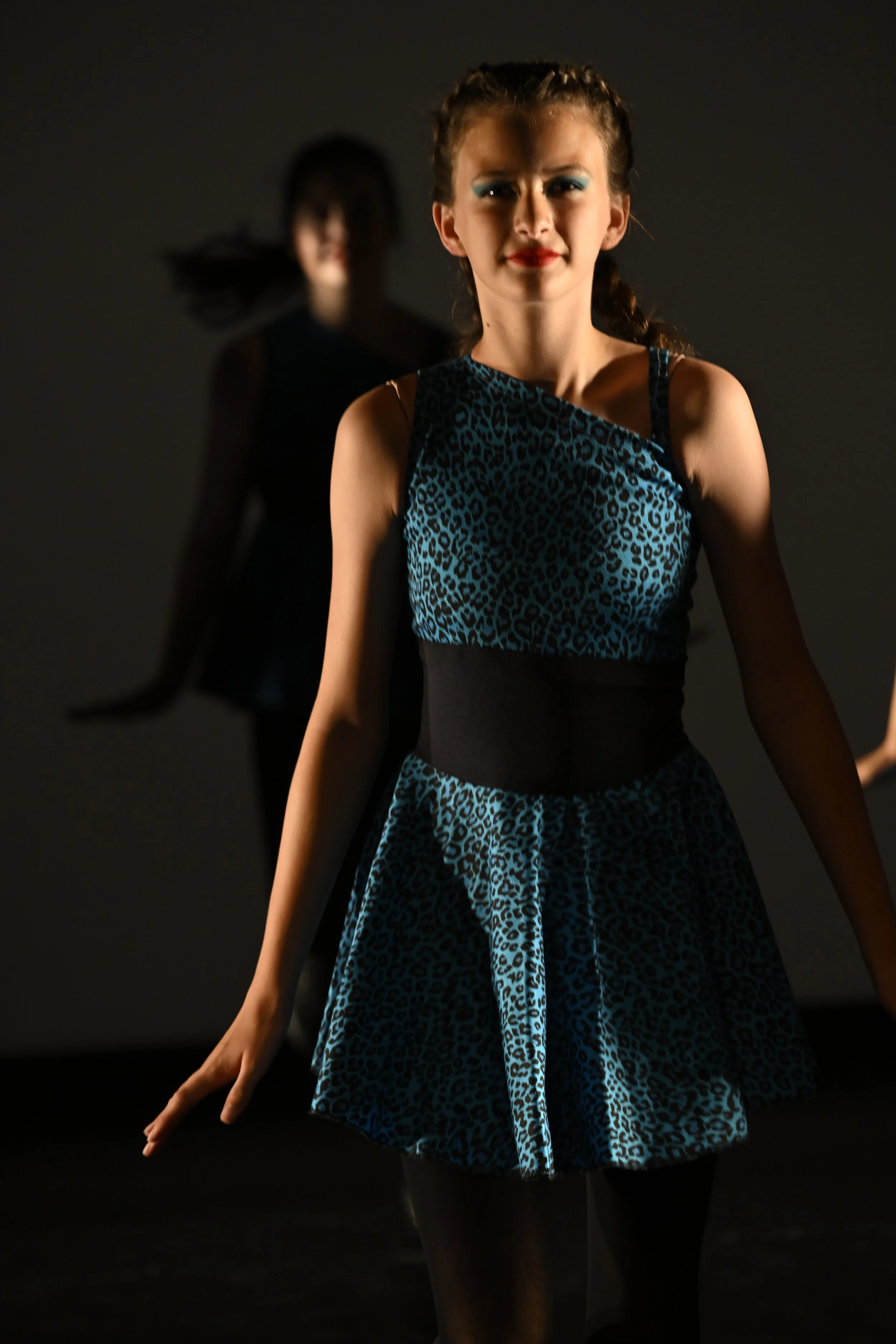 Two young women in dance costumes, one in the foreground wearing a blue leopard print dress with a black waistband, and the other in the background wearing a dark dress, both in a dark setting, possibly a dance studio or performance space.