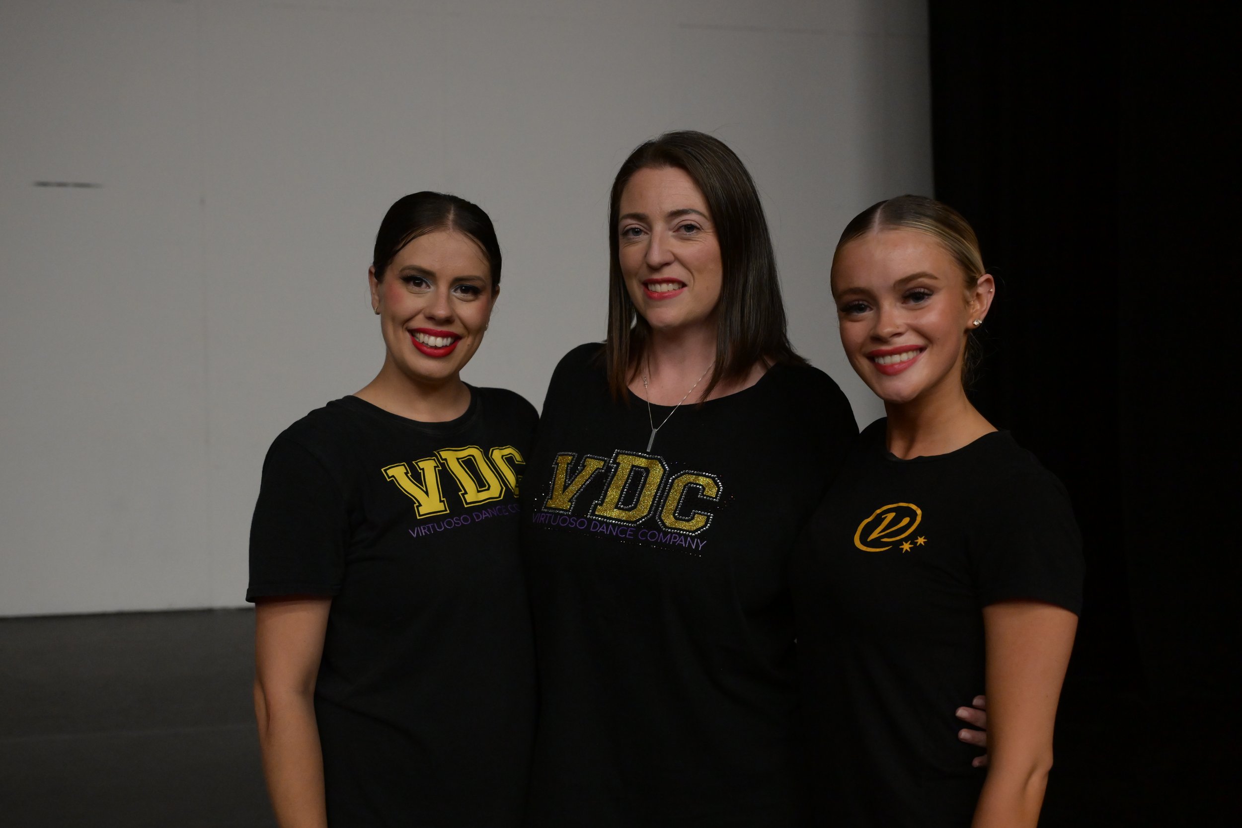 Three women standing side by side, smiling, wearing black t-shirts with the letters "VDC" and dance company logo, in front of a plain wall.