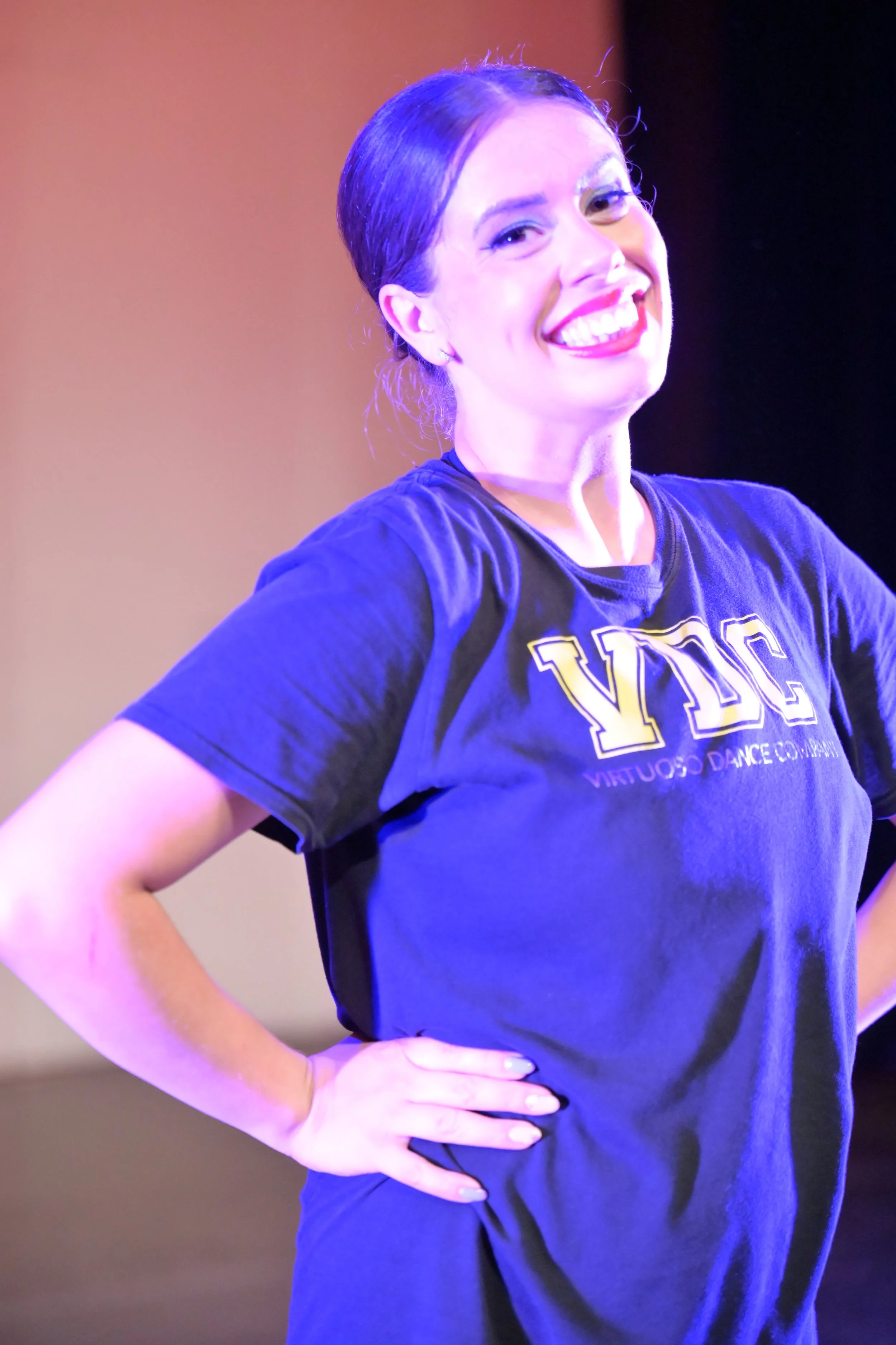 A woman with dark hair tied back, smiling confidently, wearing a black T-shirt with gold lettering and standing with her hand on her hip in front of a gradient background.