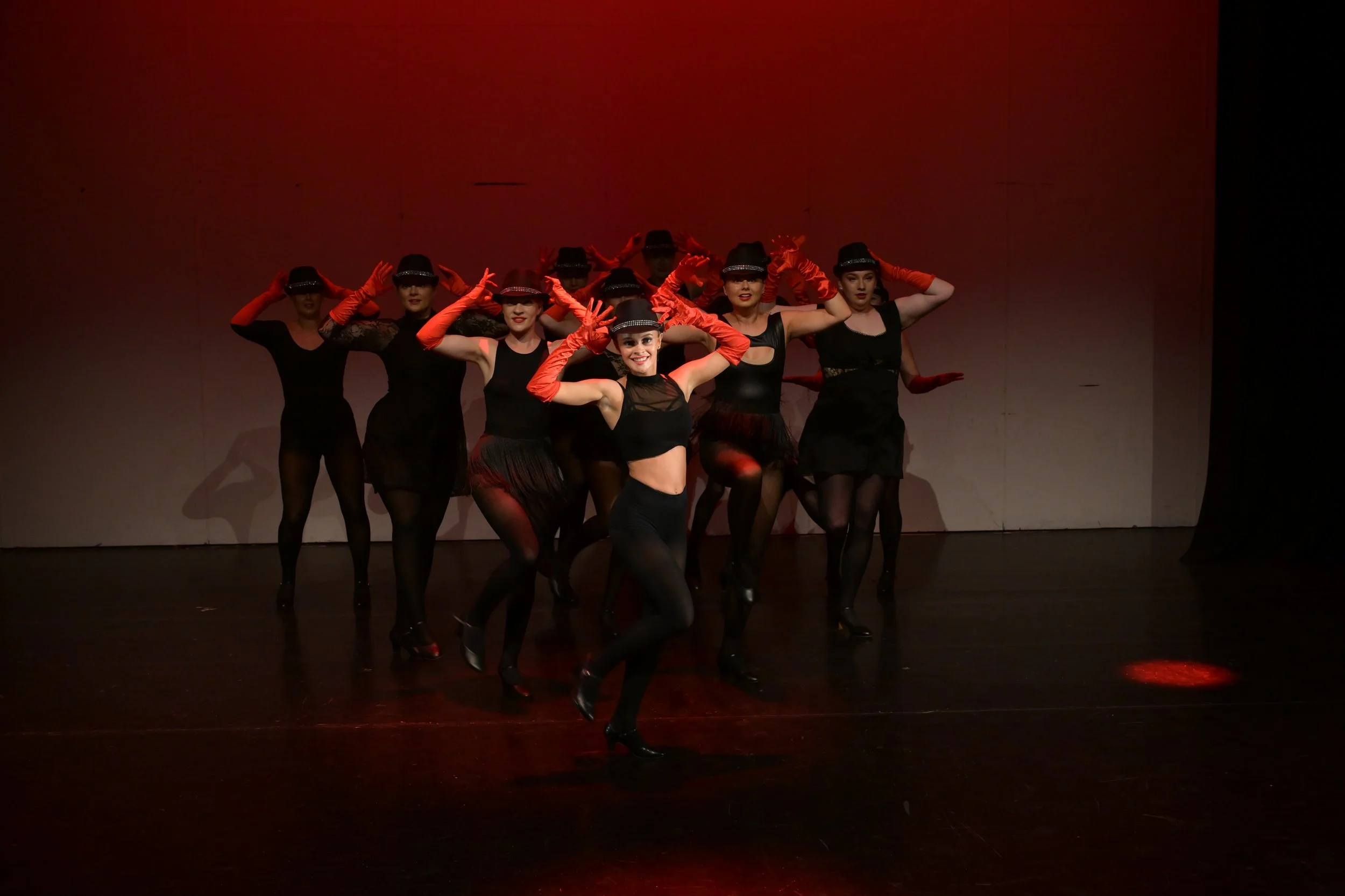 A group of female performers dressed in black dance costumes, wearing black hats, and red gloves on stage