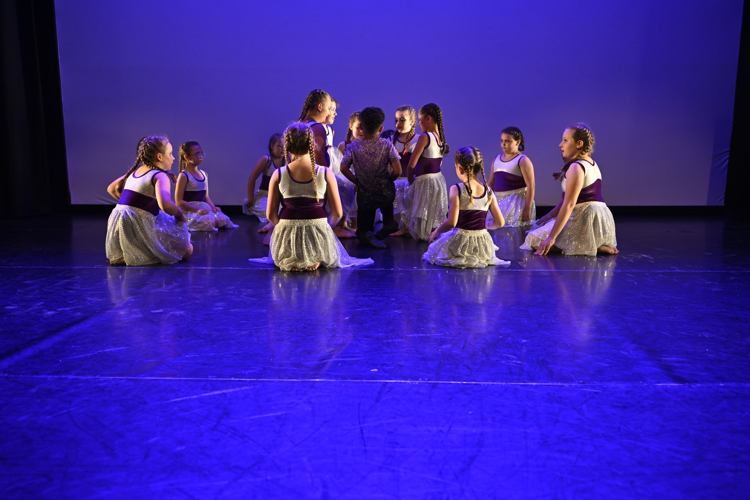 Group of young girls in ballet costumes kneeling on stage, with two children standing in the center, under blue stage lighting.