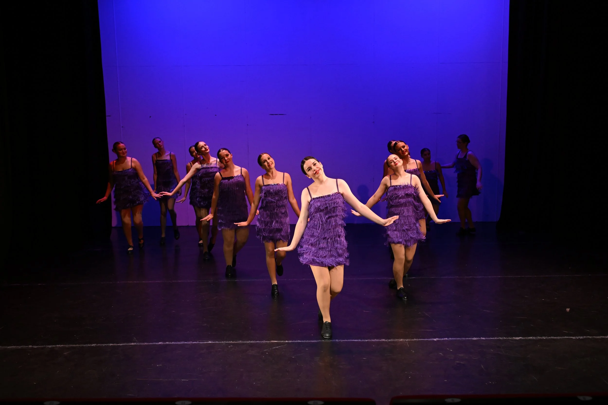 A dance troupe performing on stage, wearing purple fringed dresses, with coordinated dance poses under purple stage lighting.