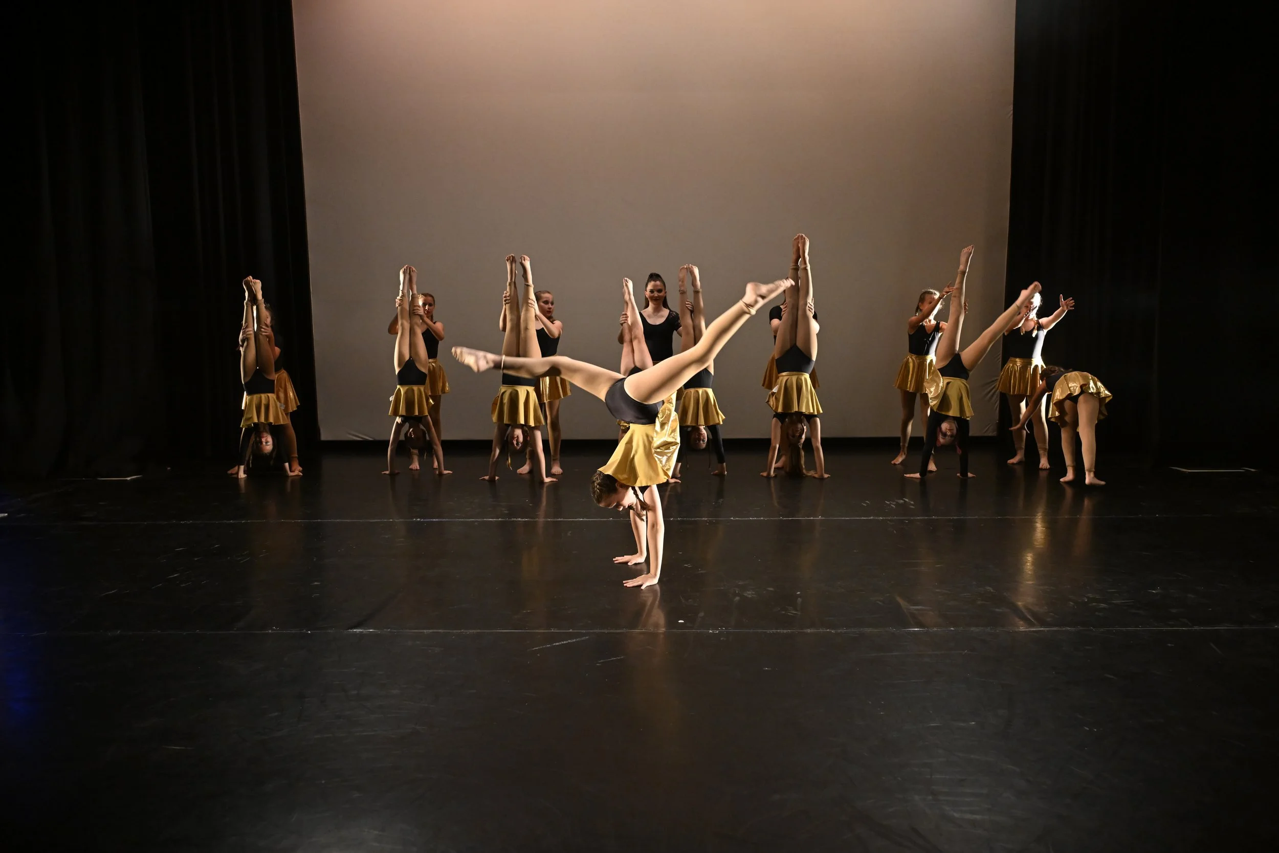 A group of young dancers performing on stage, with one girl in the front doing a handstand with legs extended outward, dressed in black and gold costumes.