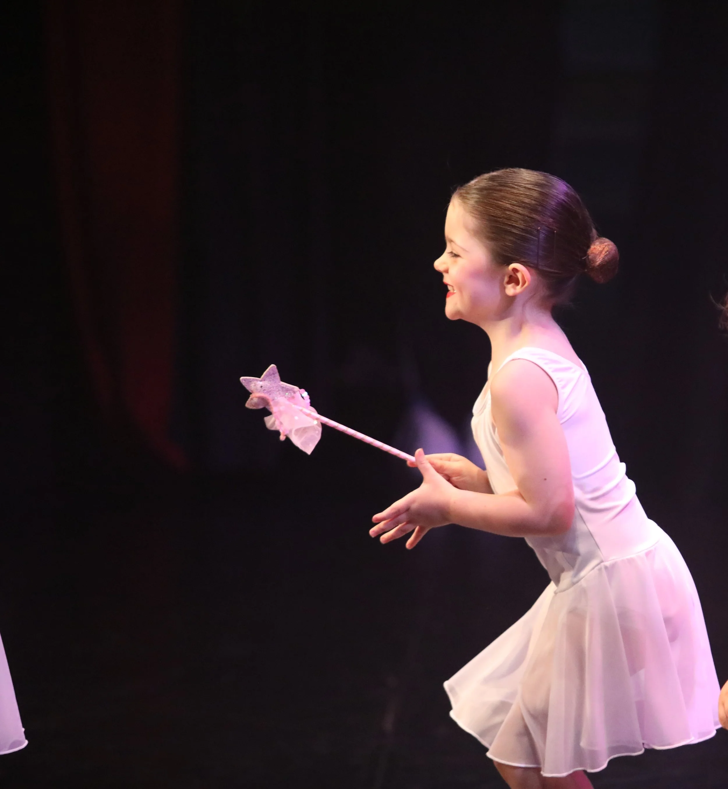 A young girl in a white dress holding a pink star-shaped magic wand, with her hair in a bun, smiling as she dances on stage.