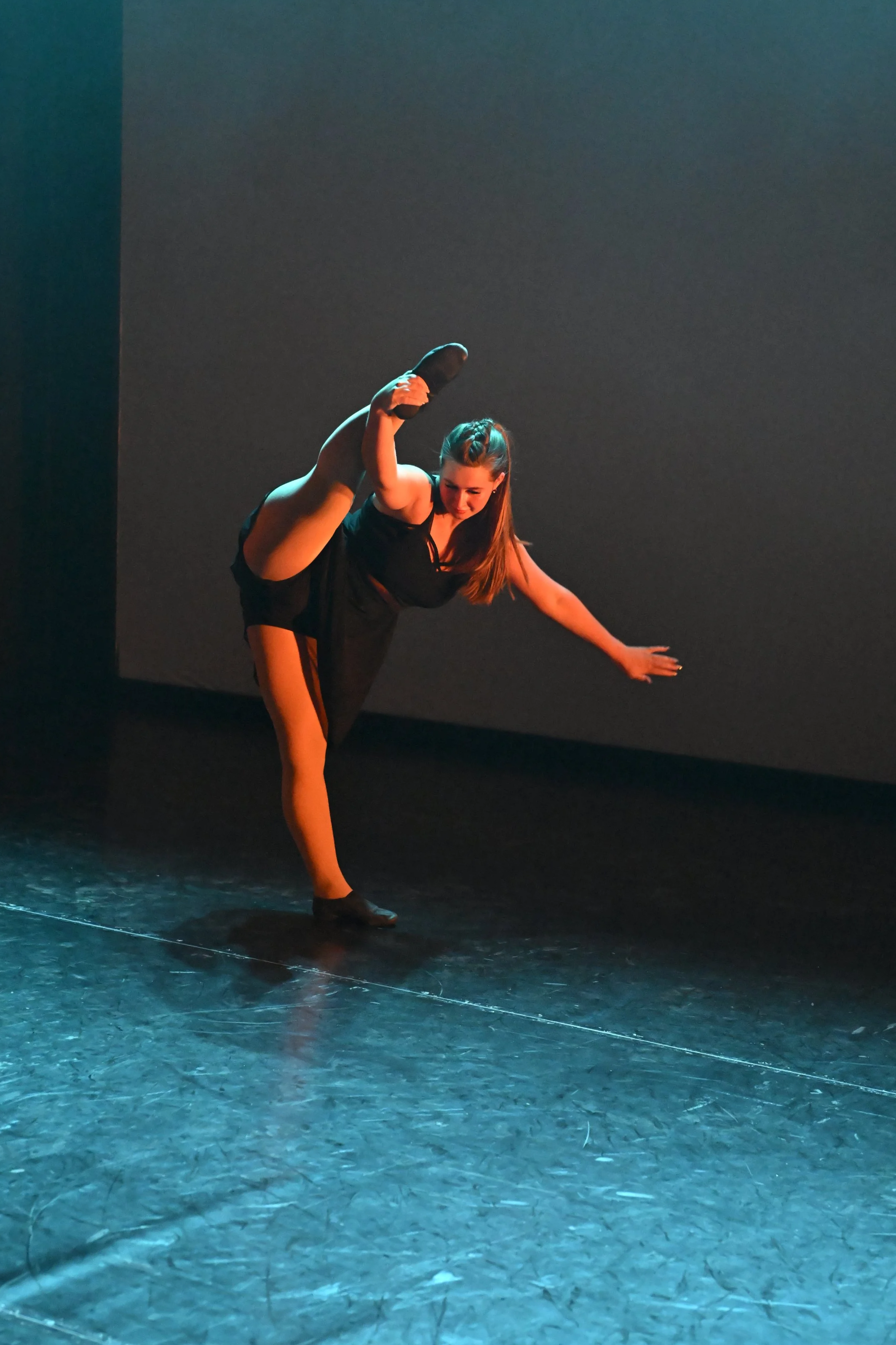 A female dancer performing on stage, balancing on one leg with her other leg lifted high and out to the side, wearing a black dress and dance shoes.