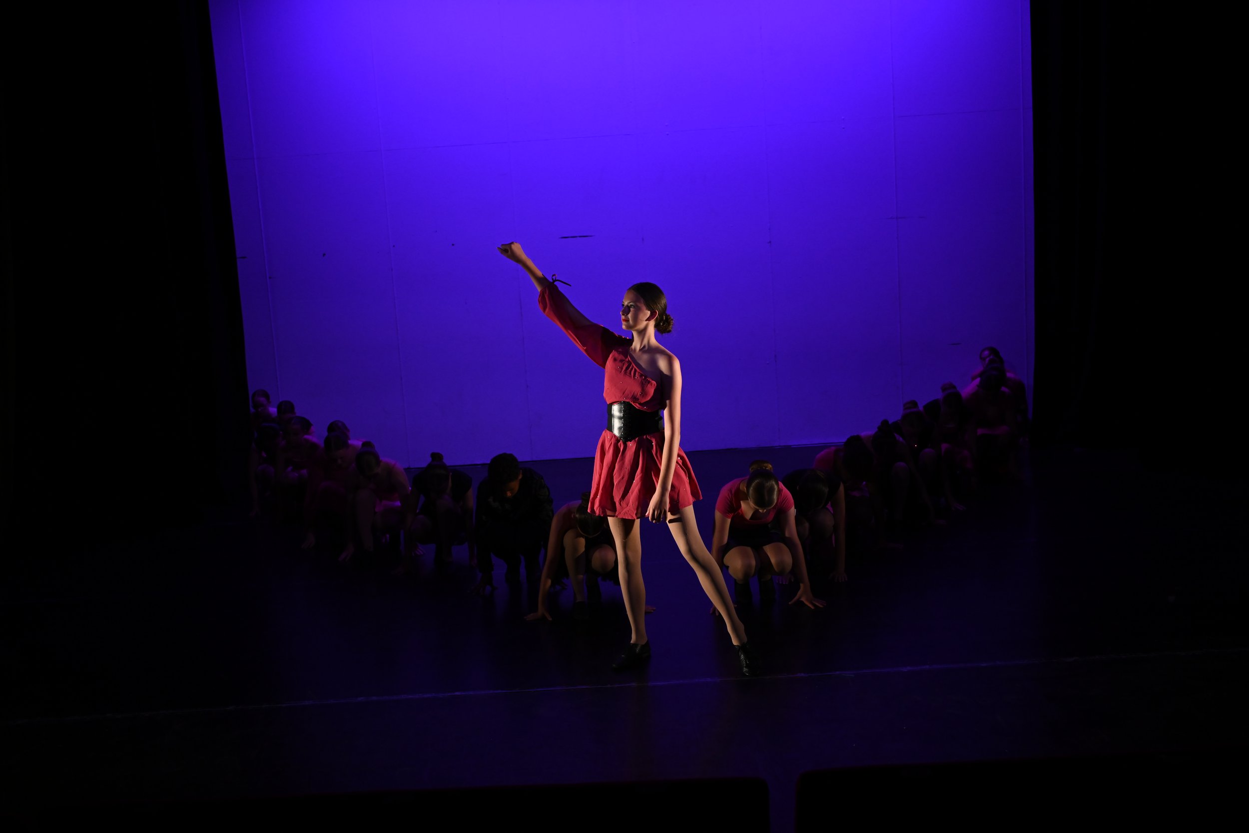 A performer in a red dress with a black belt stands with one arm raised on stage, surrounded by dancers in various poses, illuminated by purple stage lighting.