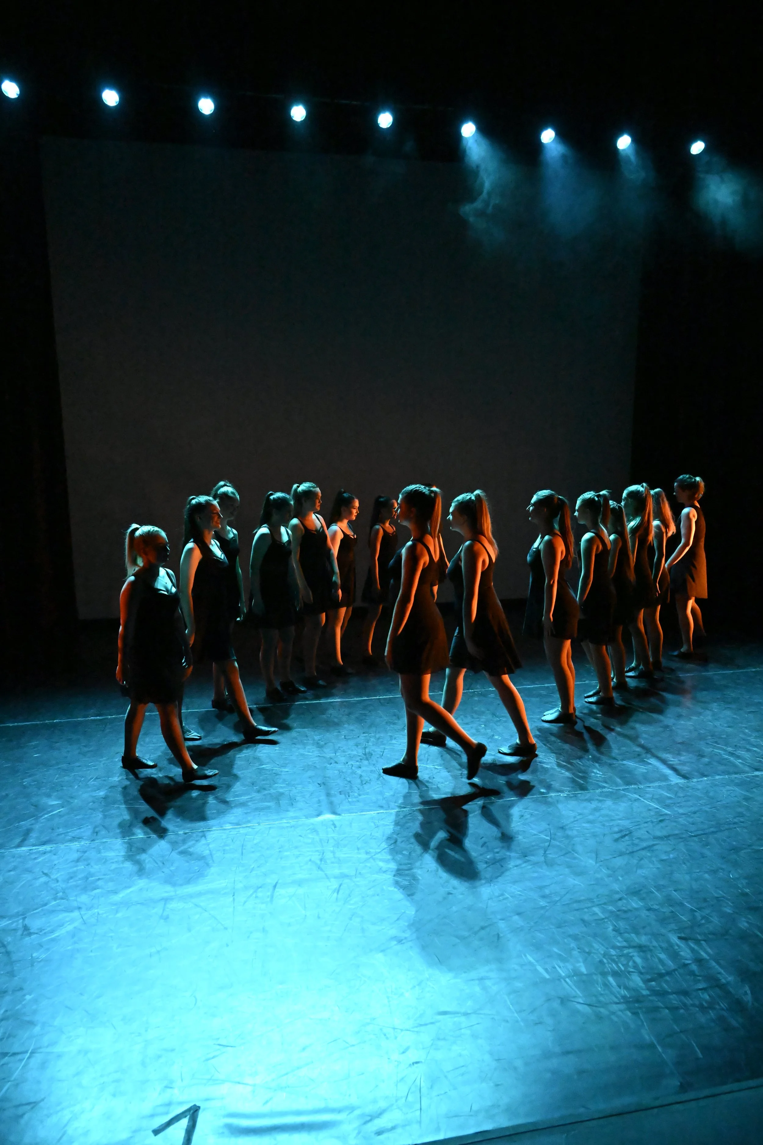 A group of young girls in black dresses performing a dance on stage with blue lighting.