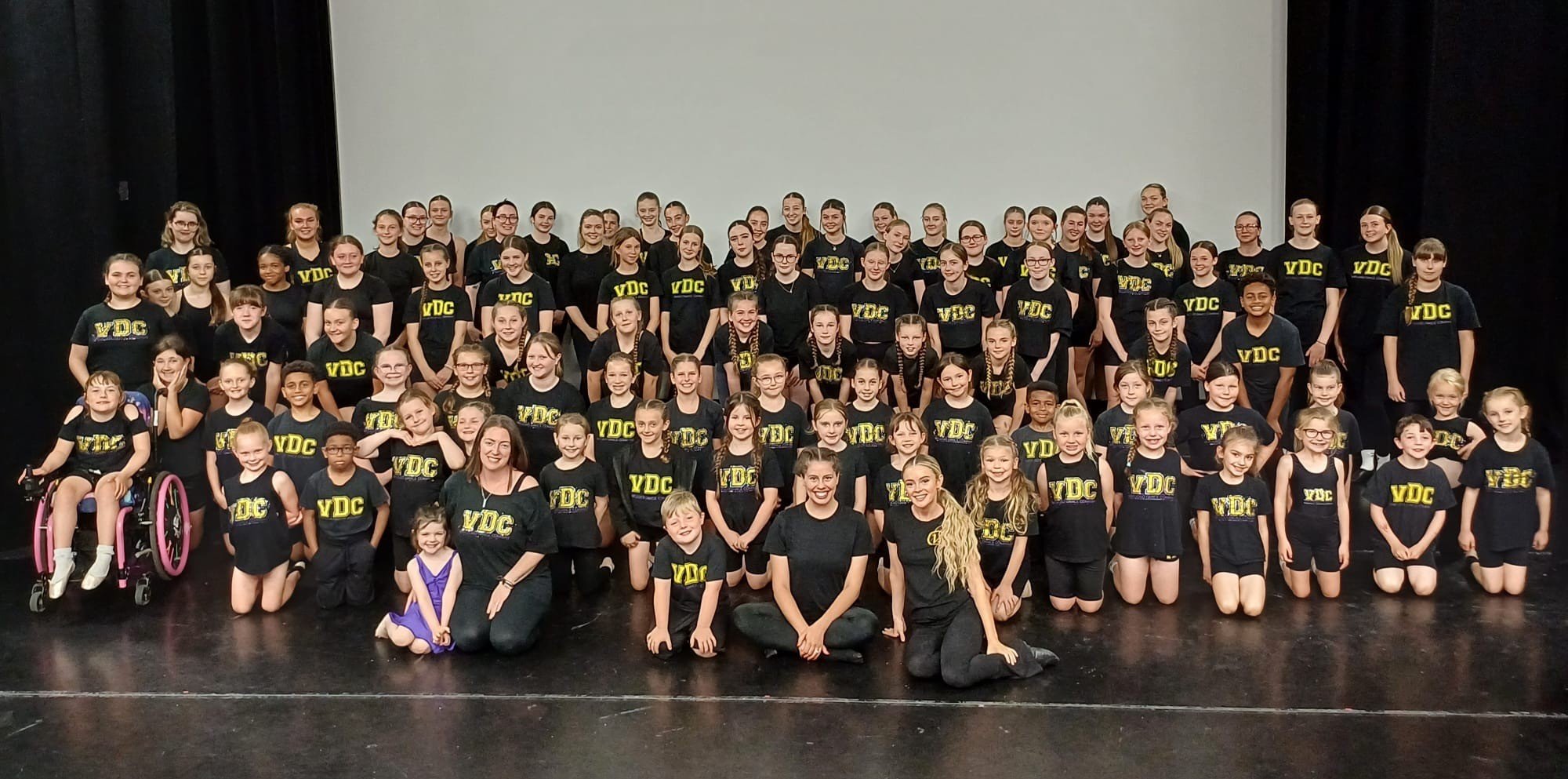 A large group of children and a few adults on a stage, wearing matching black t-shirts with yellow and white text, posing for a group photo.