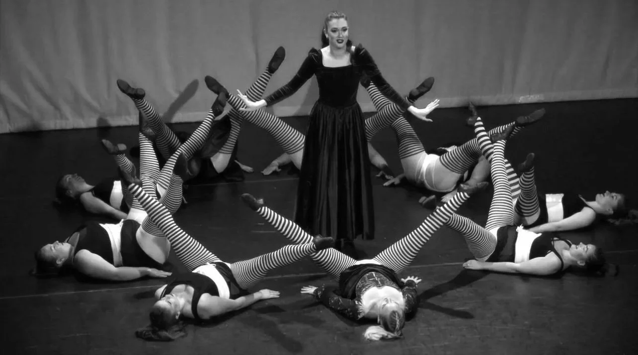 A woman in a long dress stands with her arms outstretched, surrounded by eight women in striped tights and dance costumes lying on the floor in various poses.