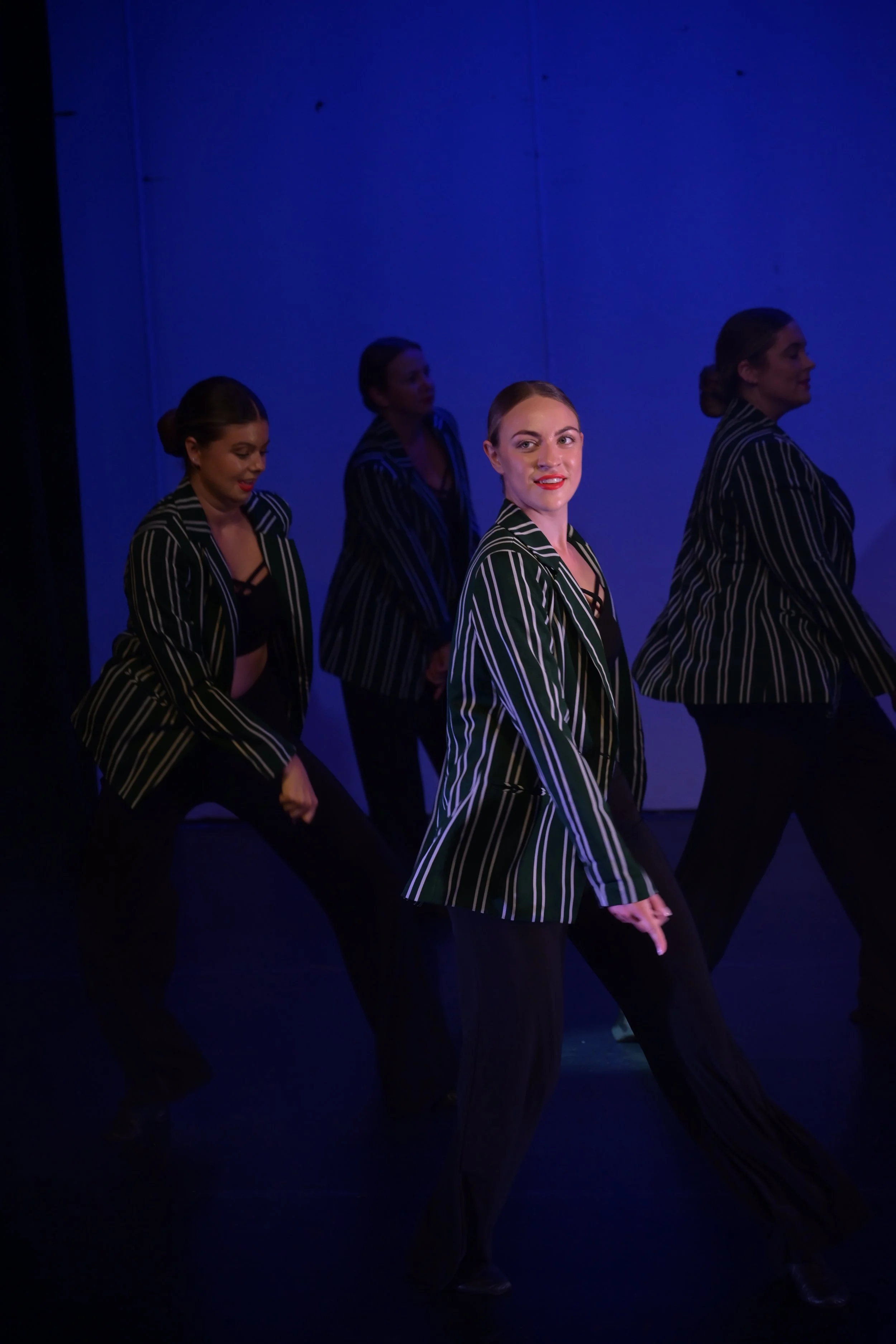 Four women in striped blazers performing on stage with a blue background.