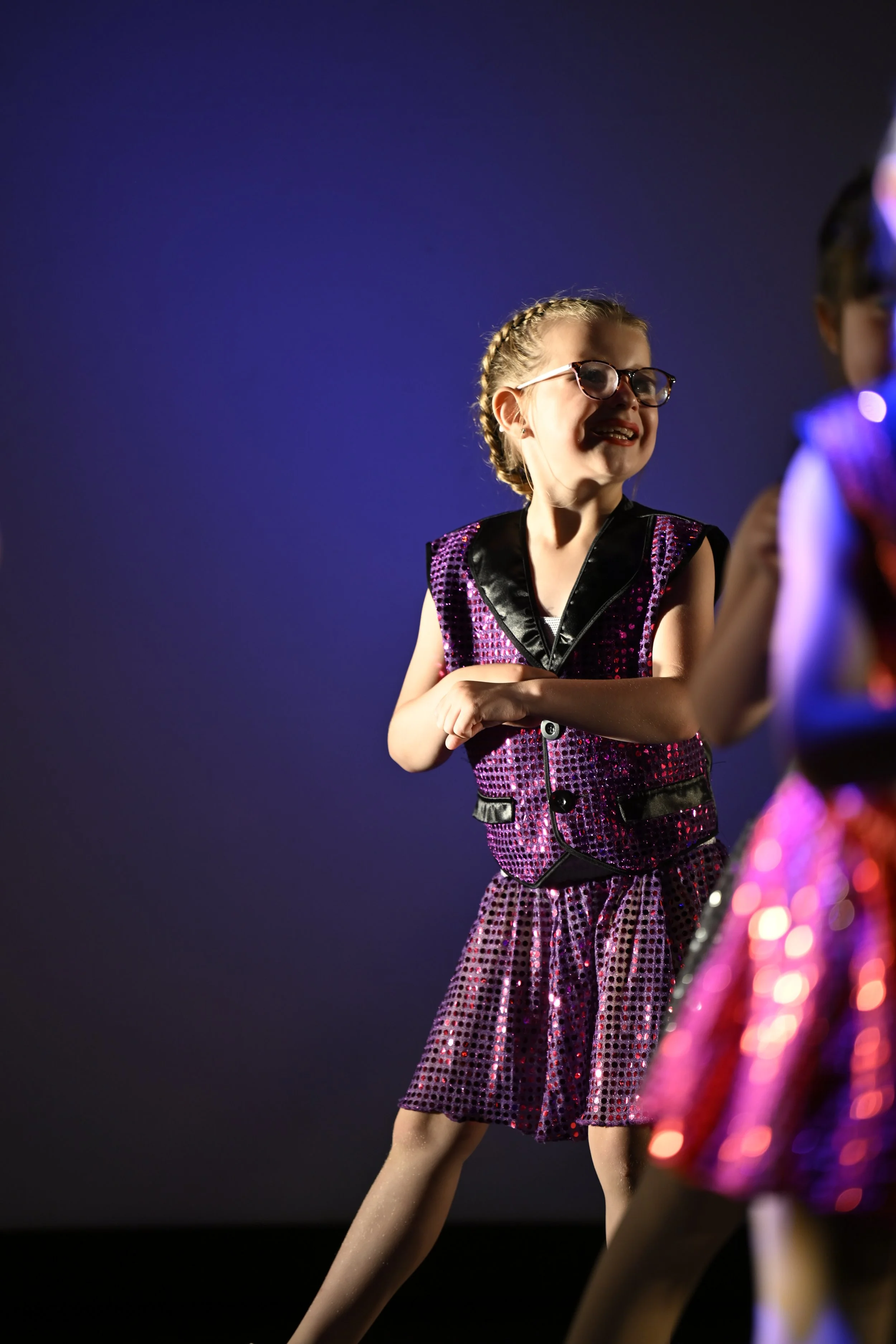 A young girl with glasses and braided hair performing on stage, wearing a shiny, pink and purple sequined costume.