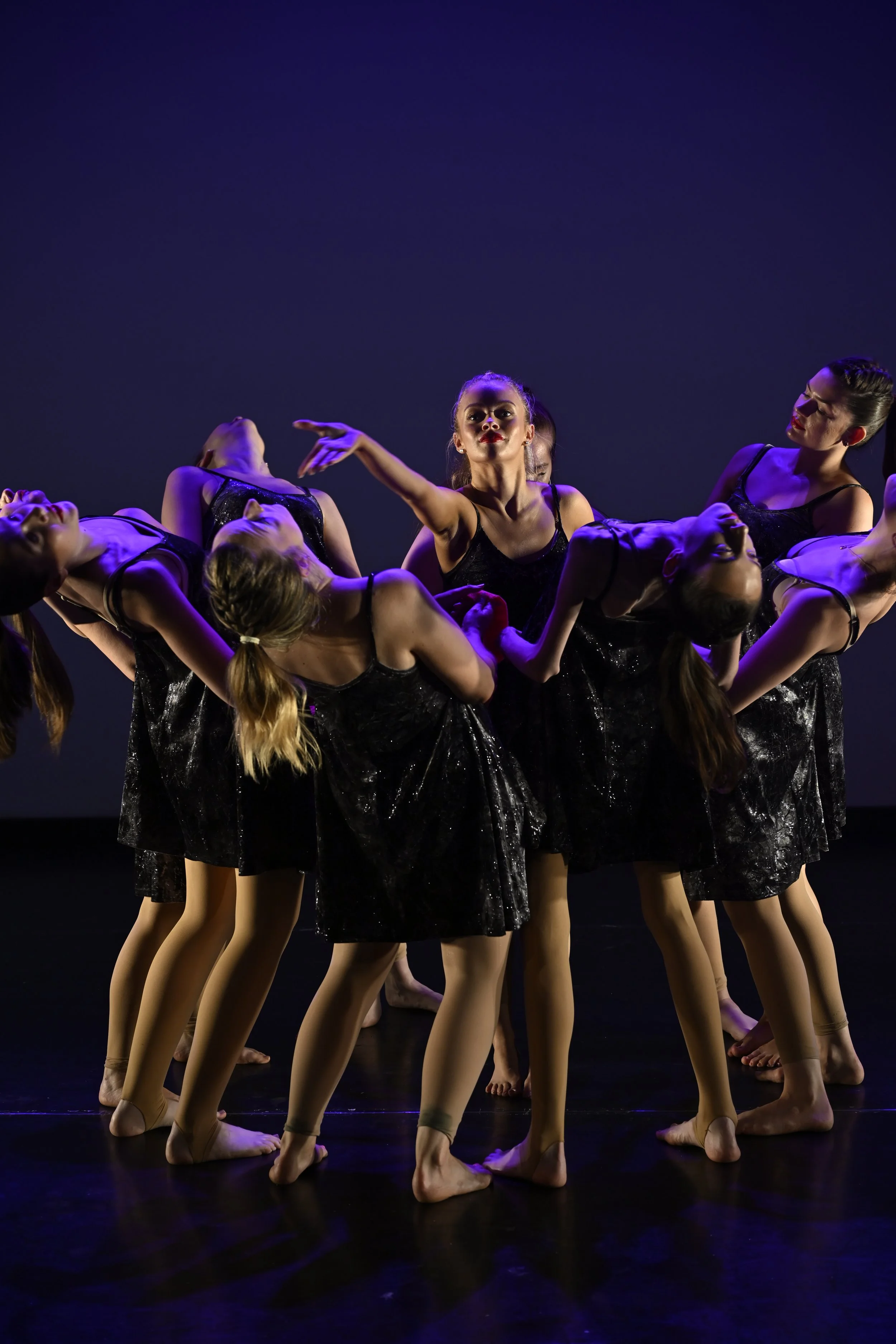Group of young female dancers in black costumes performing on stage with dark background