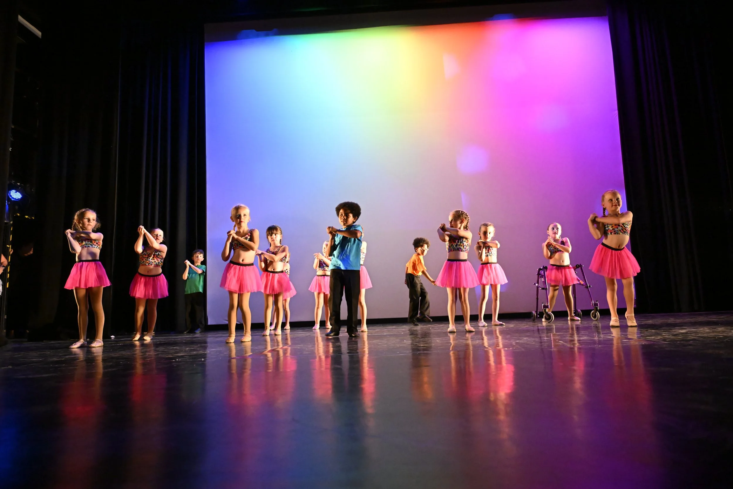 Children in colorful costumes dancing on stage during a performance with a rainbow-colored backdrop.