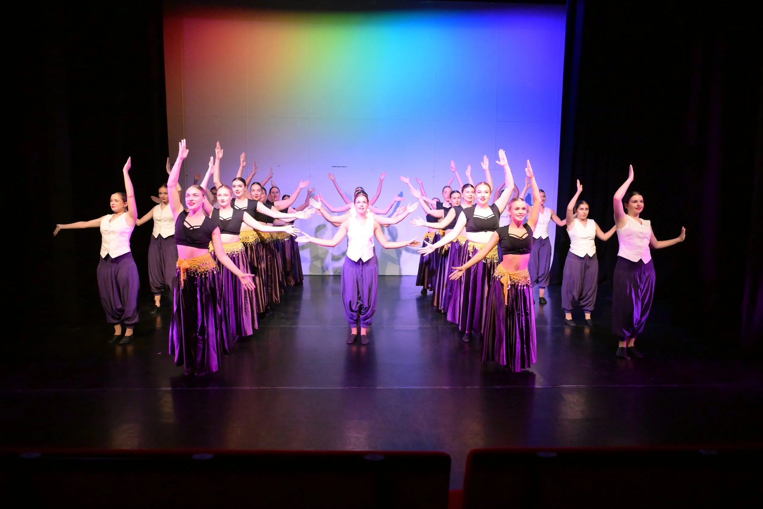 Group of female dancers performing on stage with purple skirts, white and black tops, and a colorful gradient backdrop.