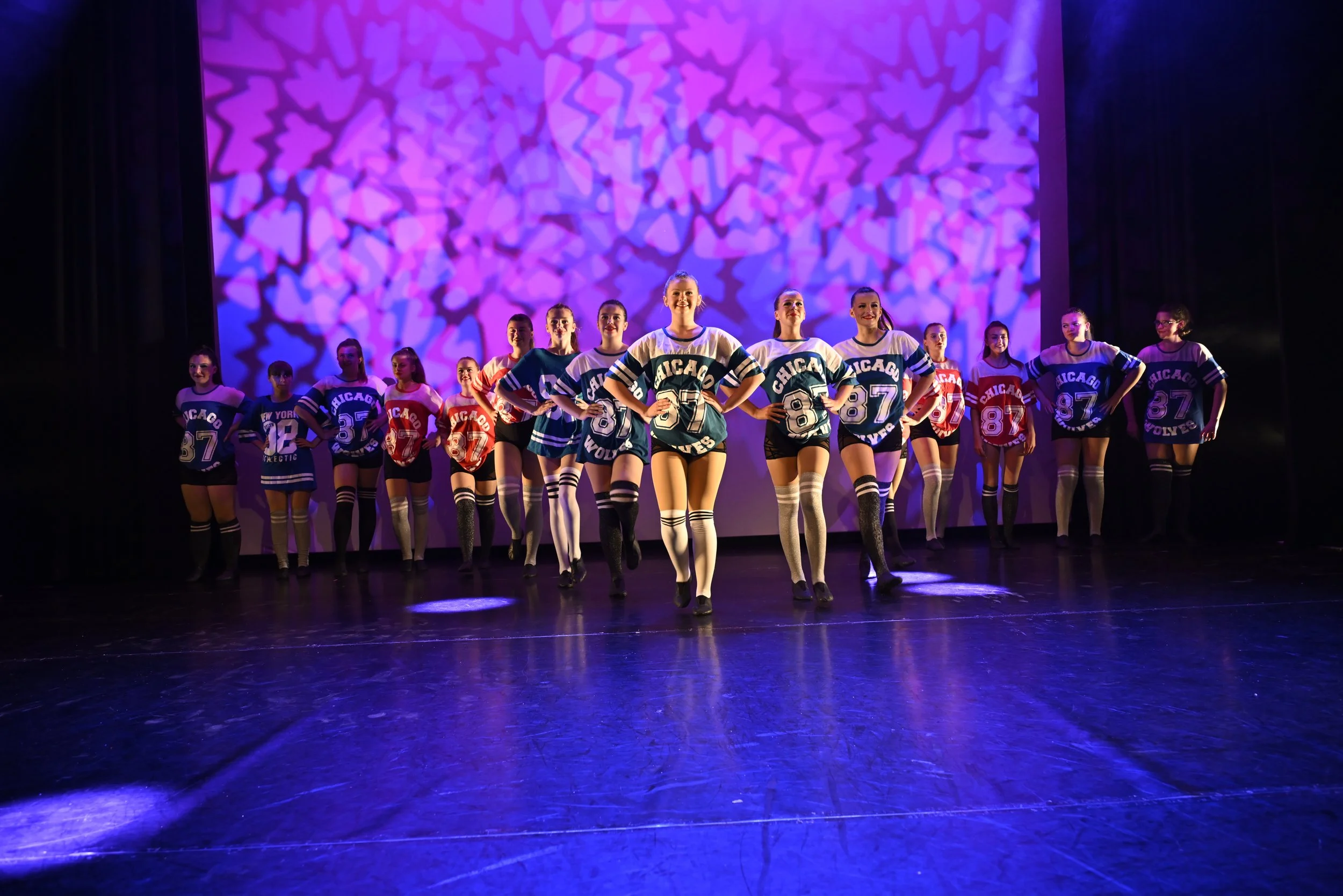 A group of dancers in sports jerseys performing on stage with a purple and pink abstract background.