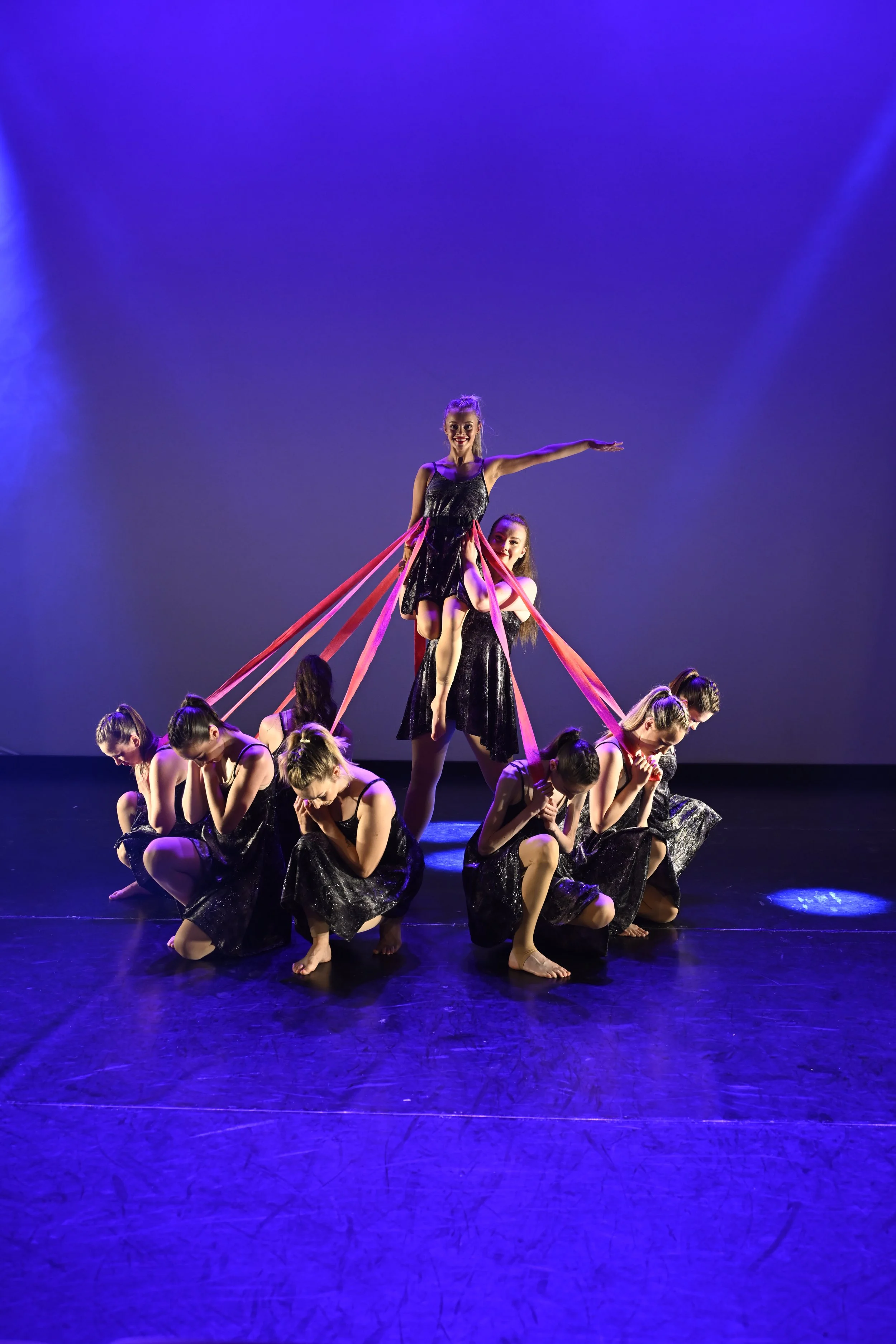 A group of young girls in black dresses performing a dance on stage with purple lighting, with some girls supporting a girl perched on their shoulders.