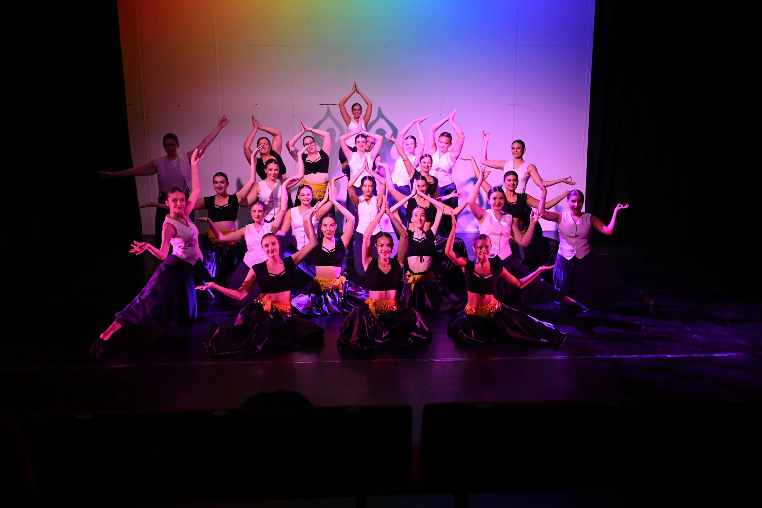 A large group of dancers performing on stage with colorful lighting and a plain backdrop.