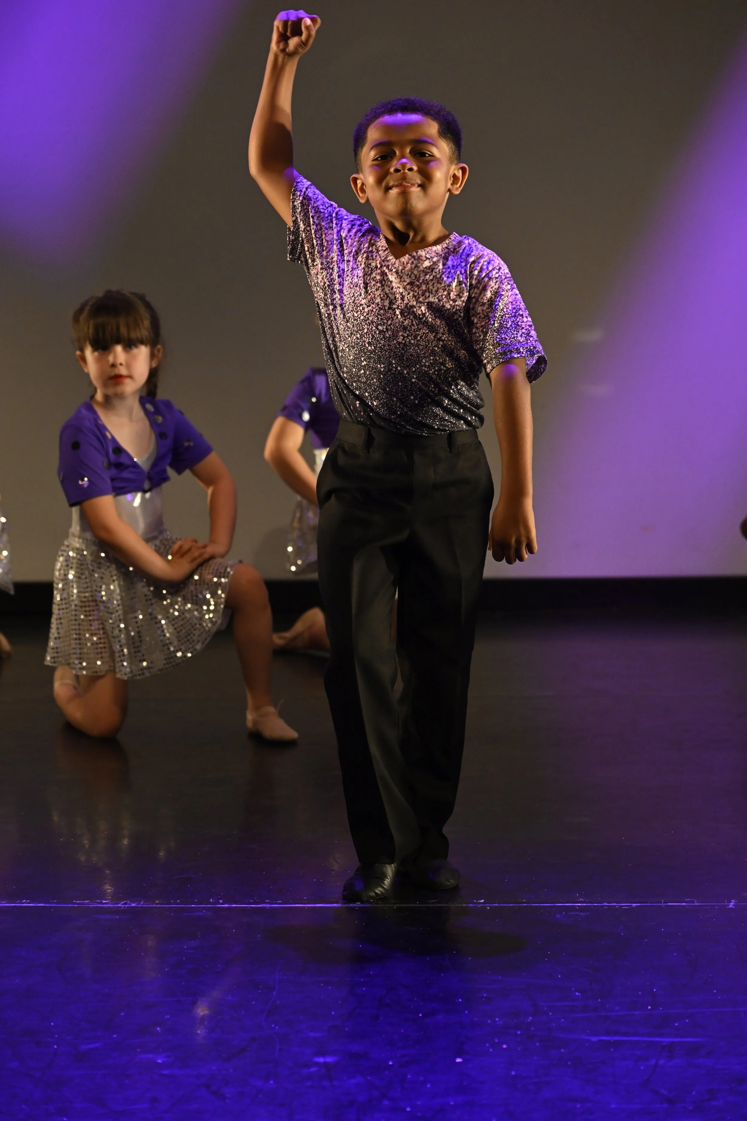 A young boy on stage performing with his arm raised, wearing a sparkly silver top and black pants, with two young girls dressed in purple tops and silver skirts in the background.